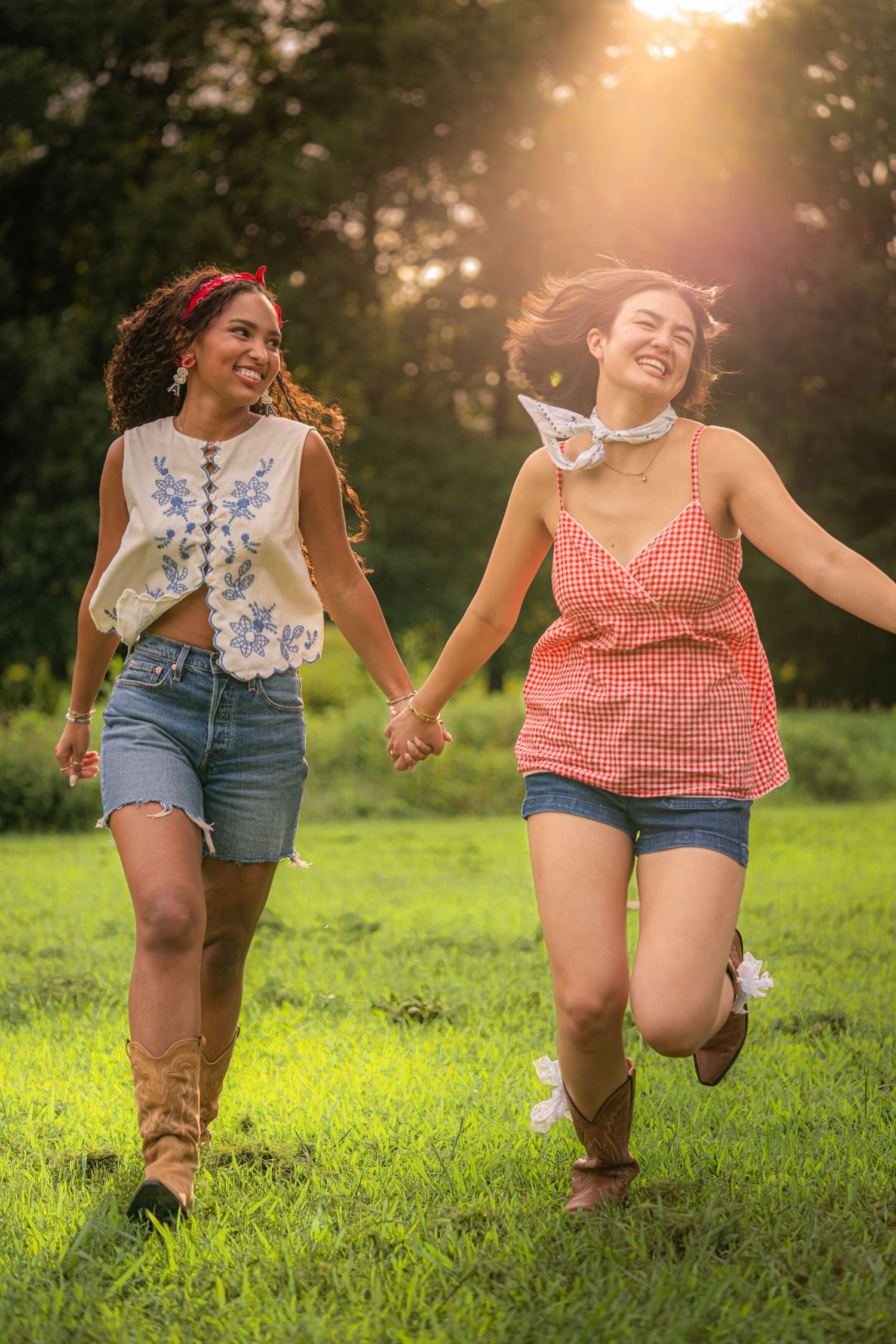 Two joyful young women holding hands and running through a sunlit green field, wearing summer outfits with denim shorts.