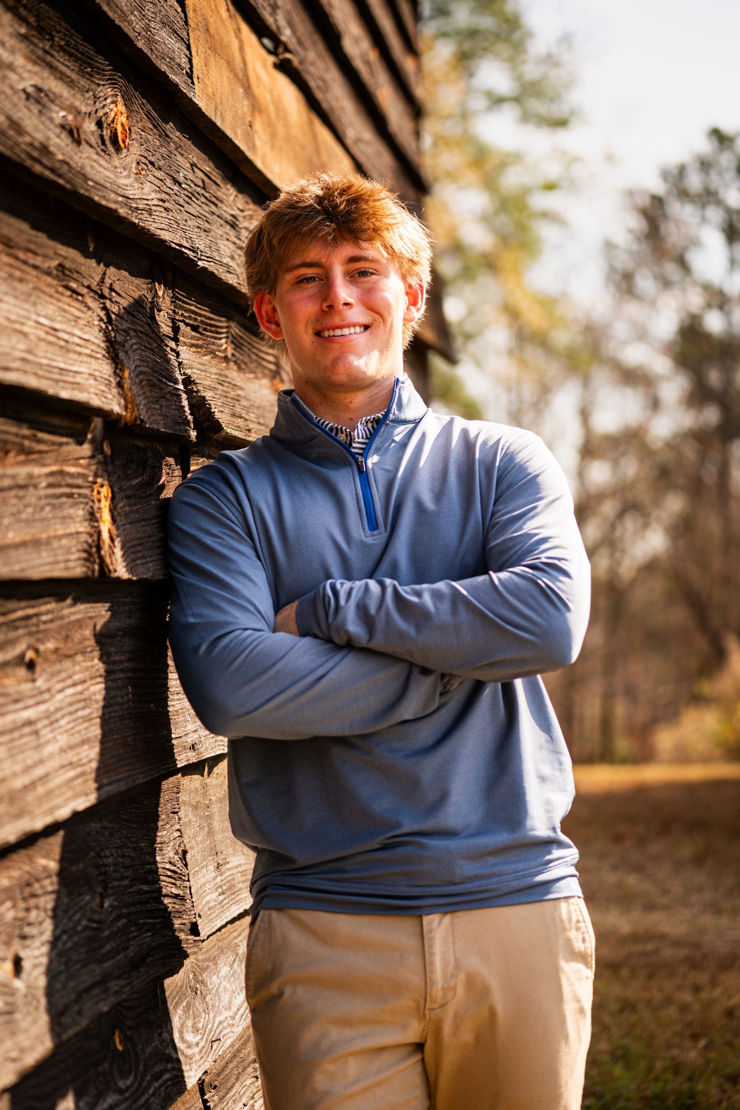 Person in a light blue quarter-zip sweater poses against rustic wooden cabin wall in warm autumn lighting.