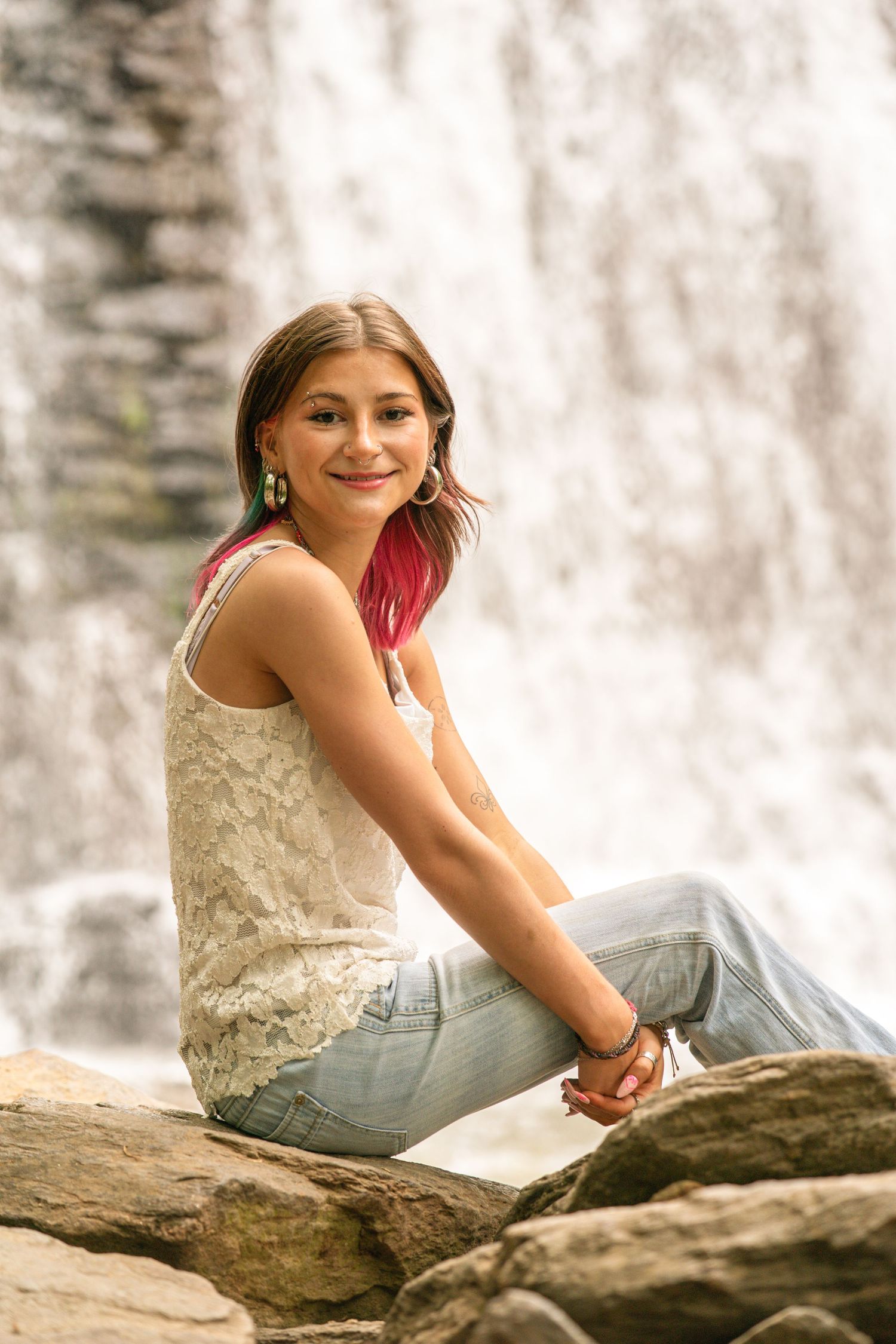 Young woman with pink highlights sits on rocks smiling before a cascading waterfall, wearing jeans and a white lace top.
