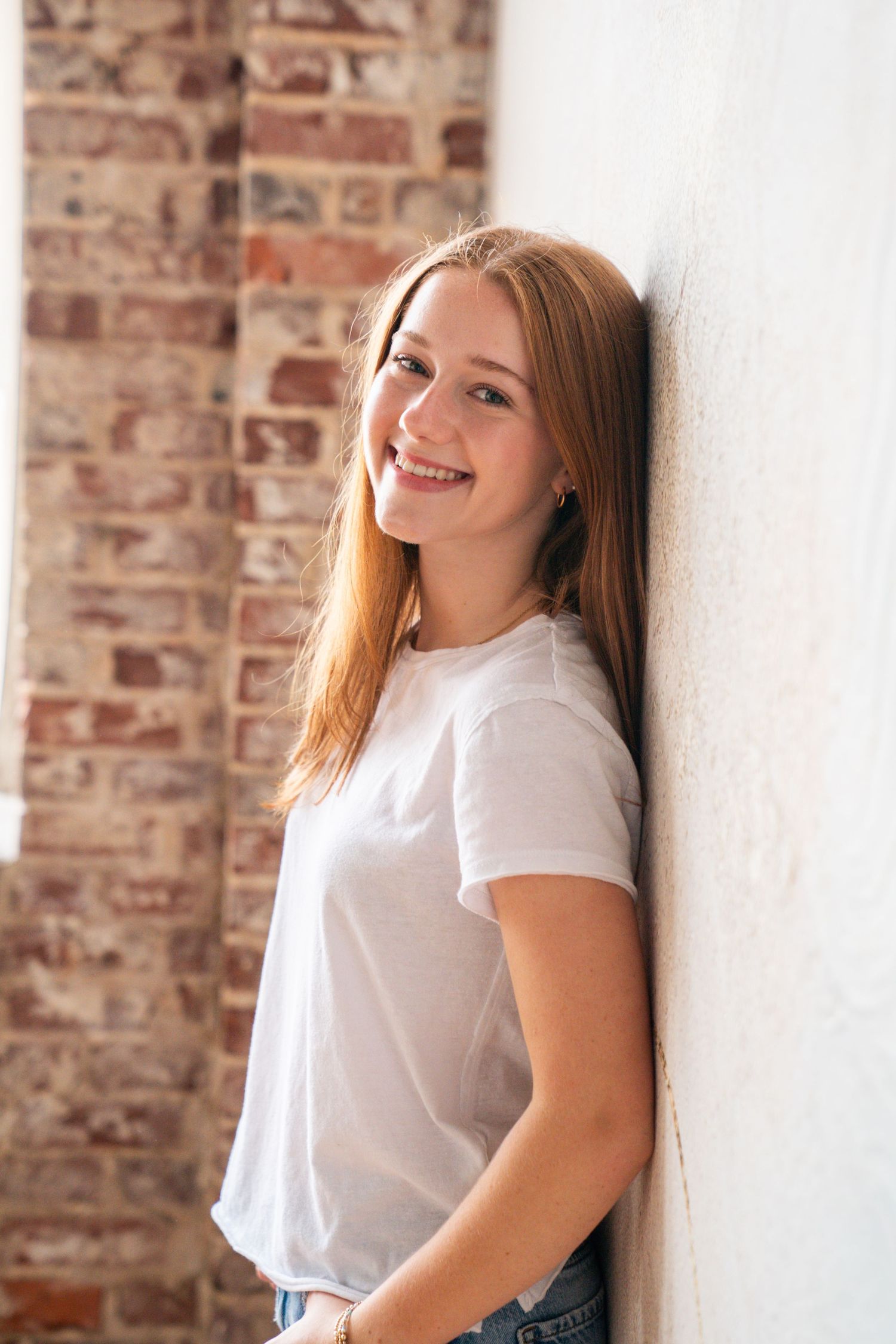 Smiling young woman with long red hair leaning against a white wall wearing a white t-shirt near a brick wall.