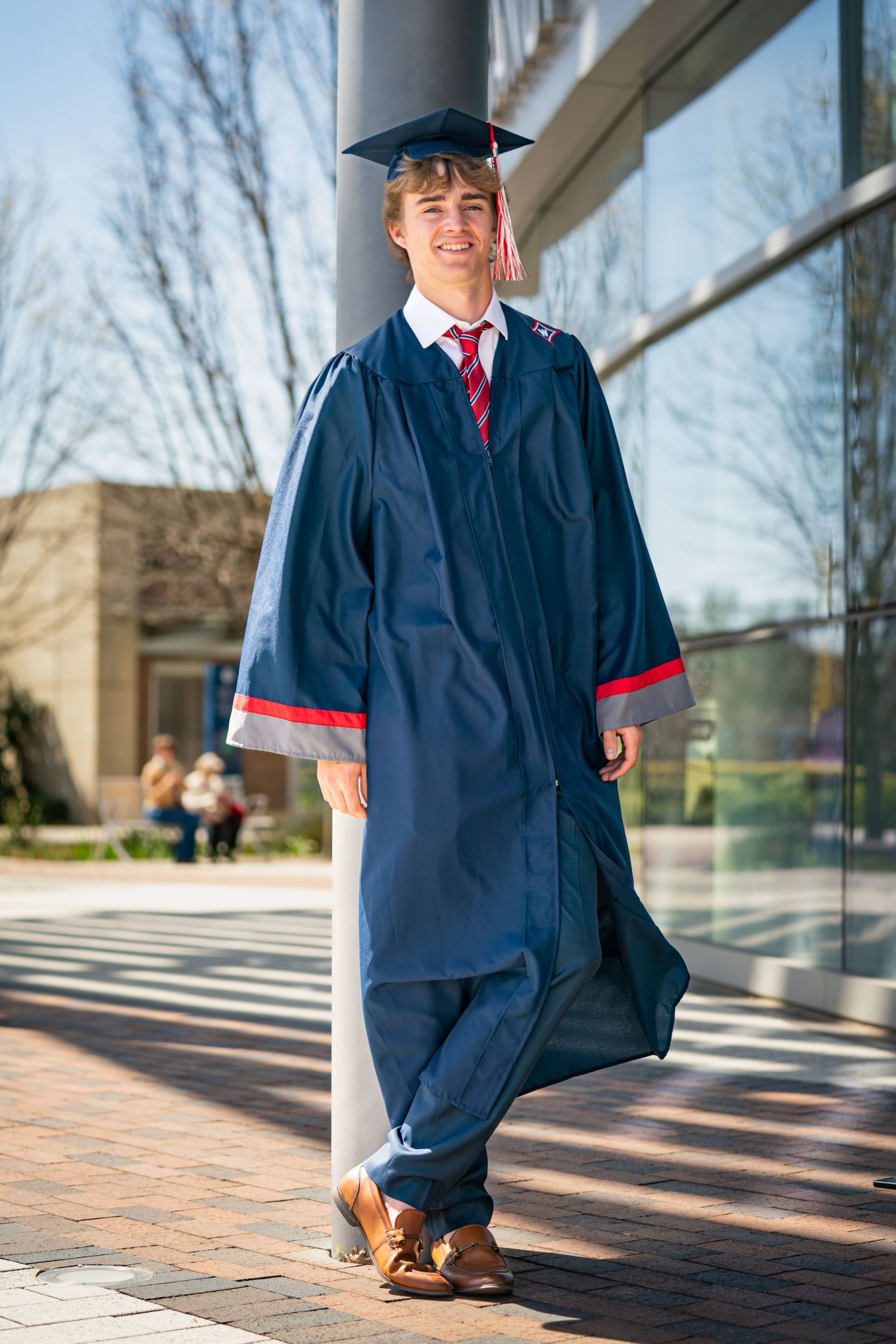Graduate in navy blue cap and gown with red tie poses outdoors against glass building on sunny day.