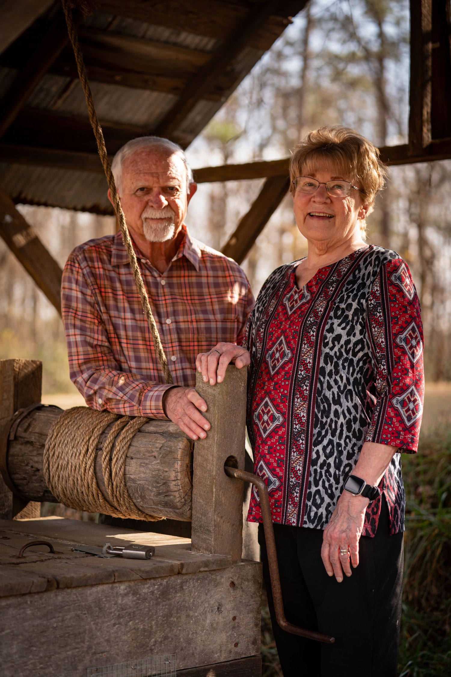 A couple in casual attire smiling and leaning on a rustic wooden fence in warm outdoor lighting.