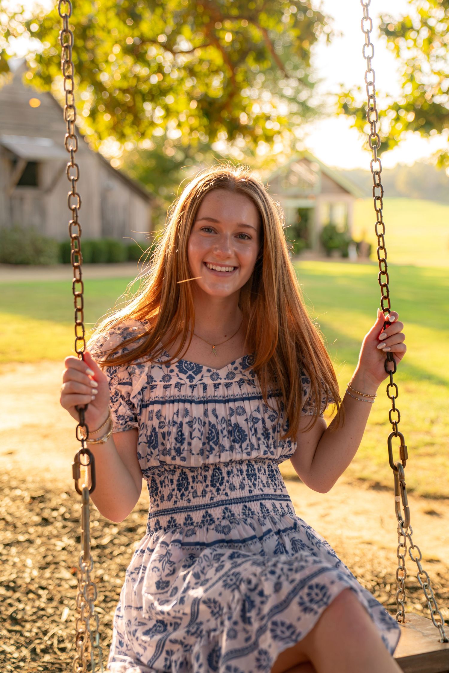 Smiling young woman with red hair in floral dress sitting on chain swing in sunny park setting.