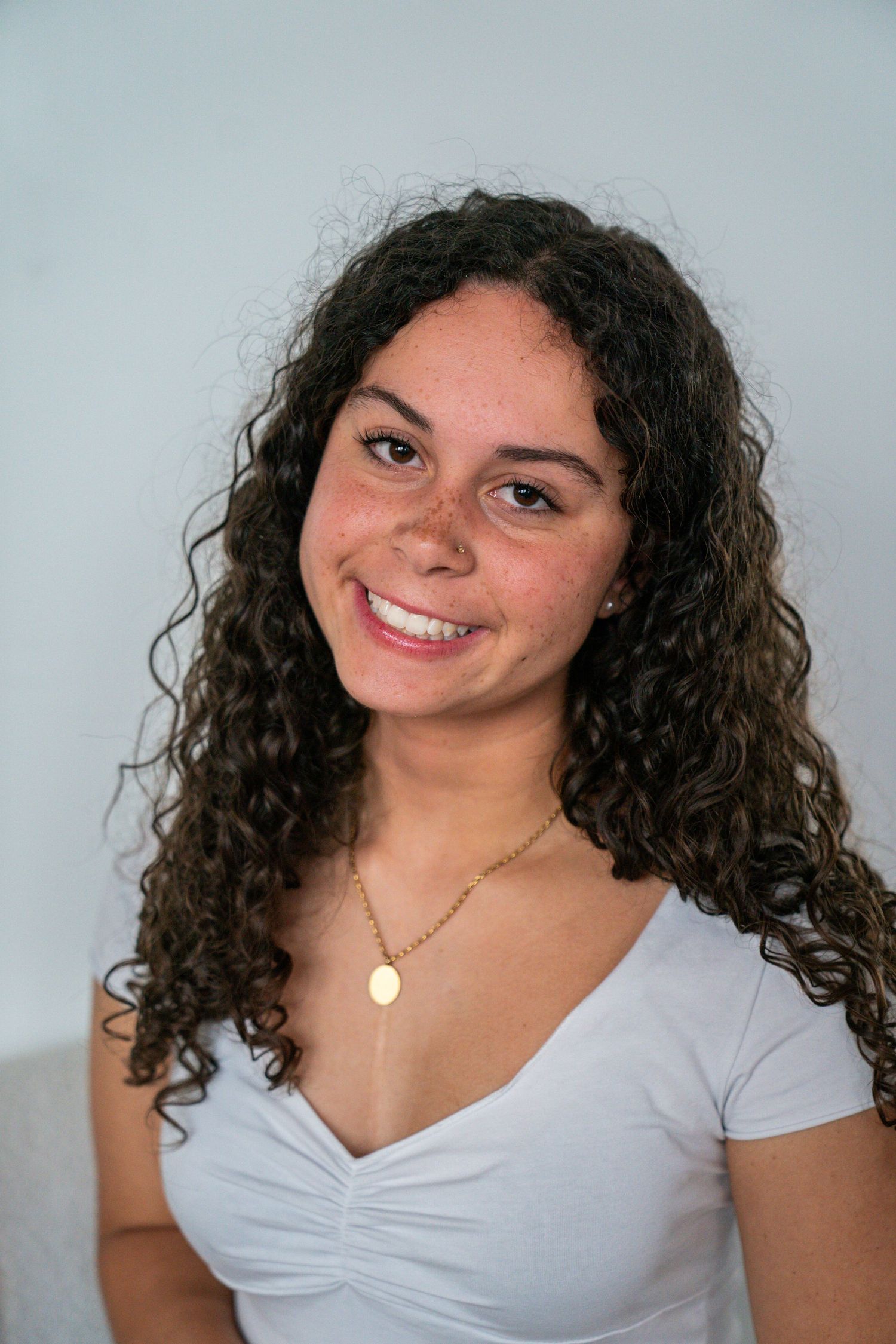 Young woman with curly hair smiling in white v-neck top, wearing gold necklace against light background.