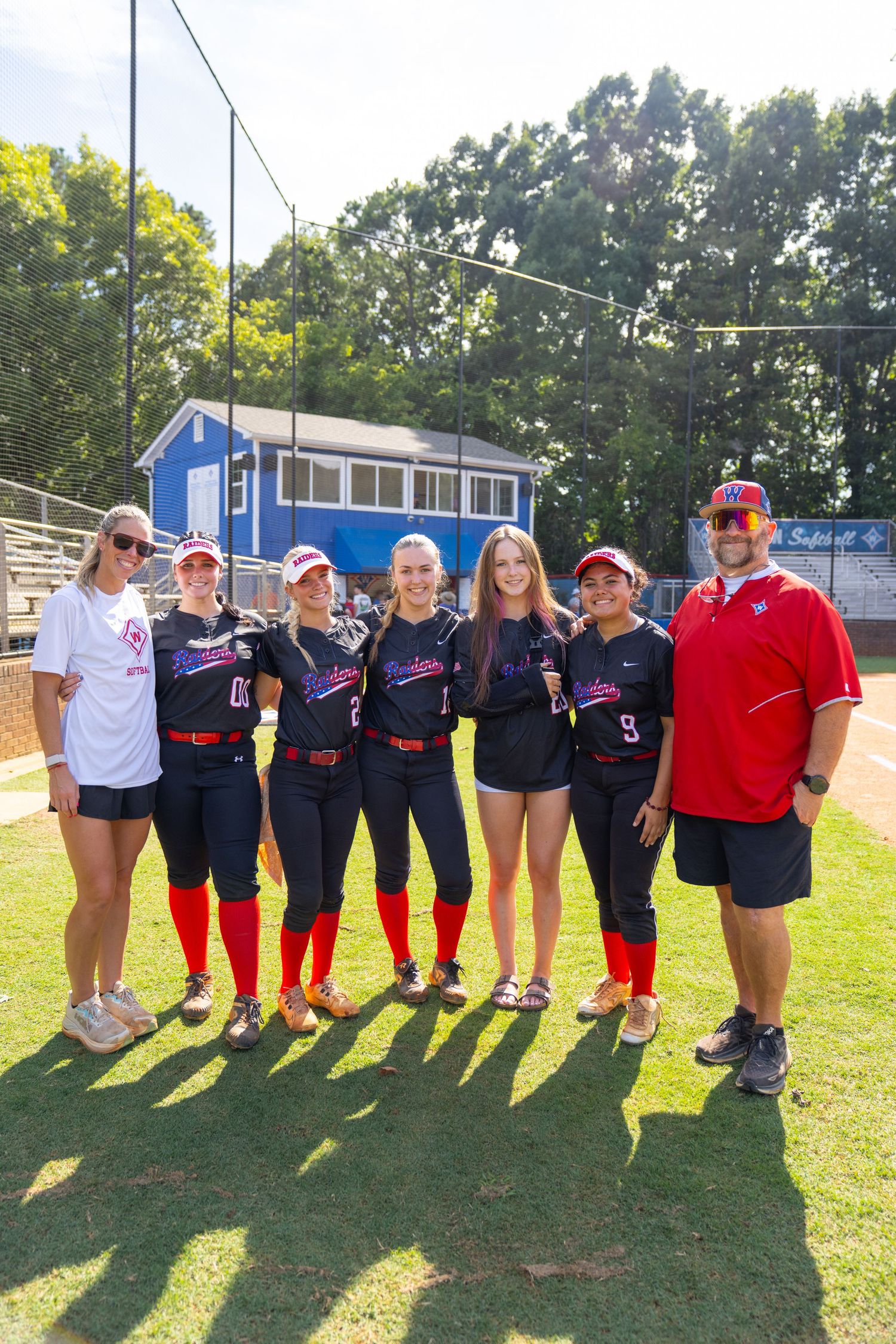Softball team poses with coaches on sunny field, wearing navy and red uniforms, with trees and blue building behind.