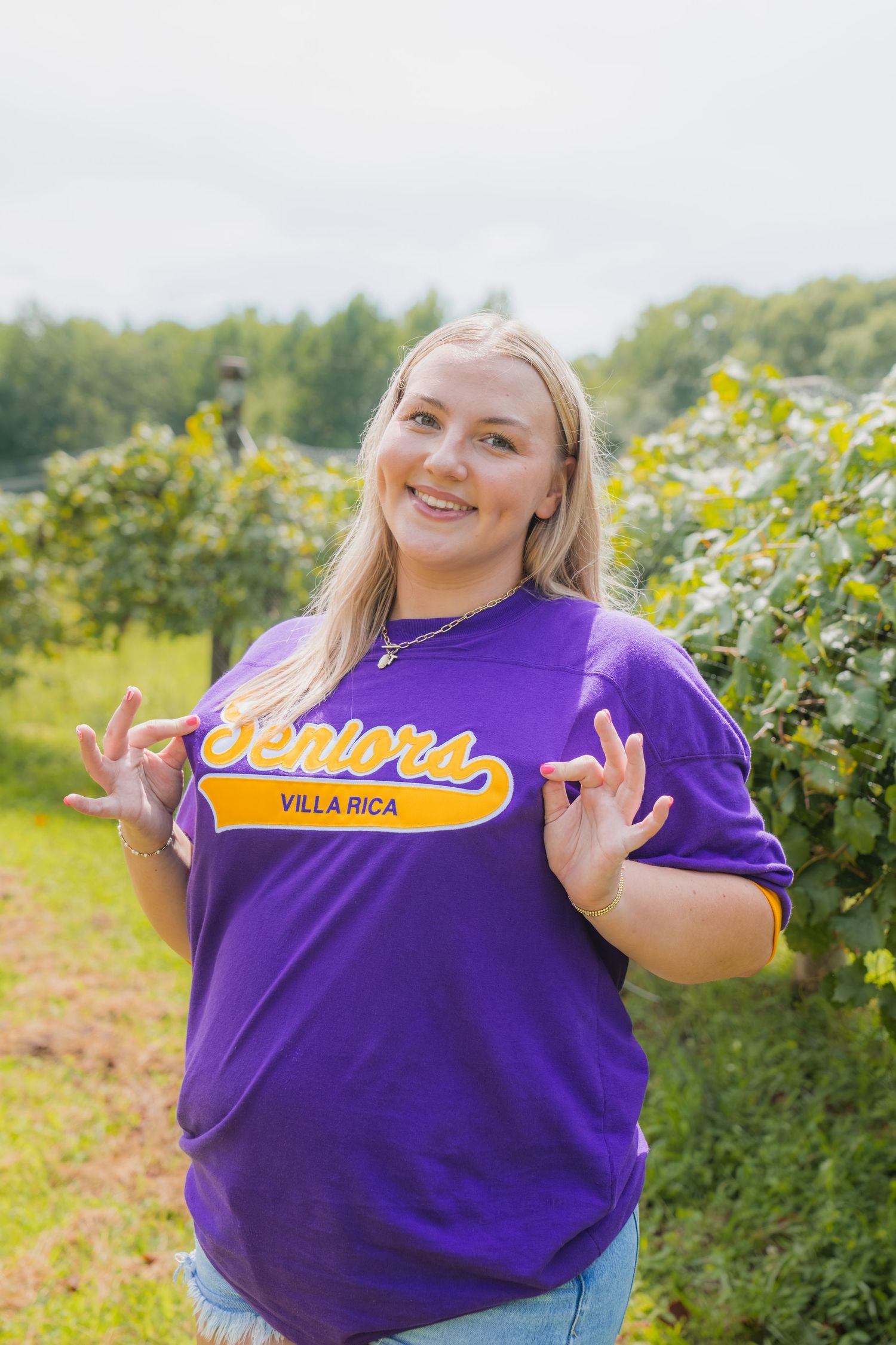 Smiling blonde woman in purple Seniors Villa Rica t-shirt posing outdoors near green vineyard rows.