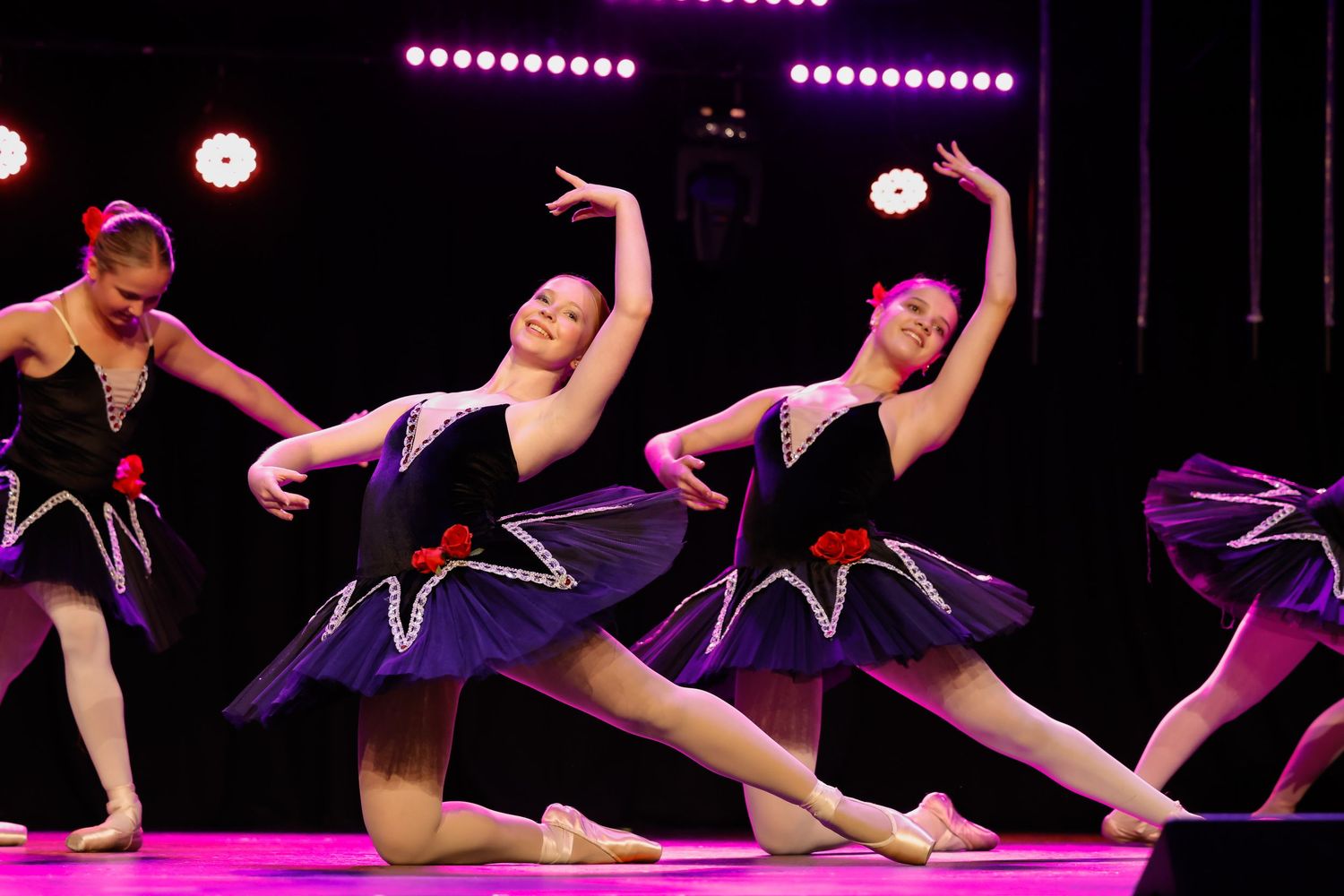 Dancers in black leotards perform a synchronized movement sequence under purple stage lighting.