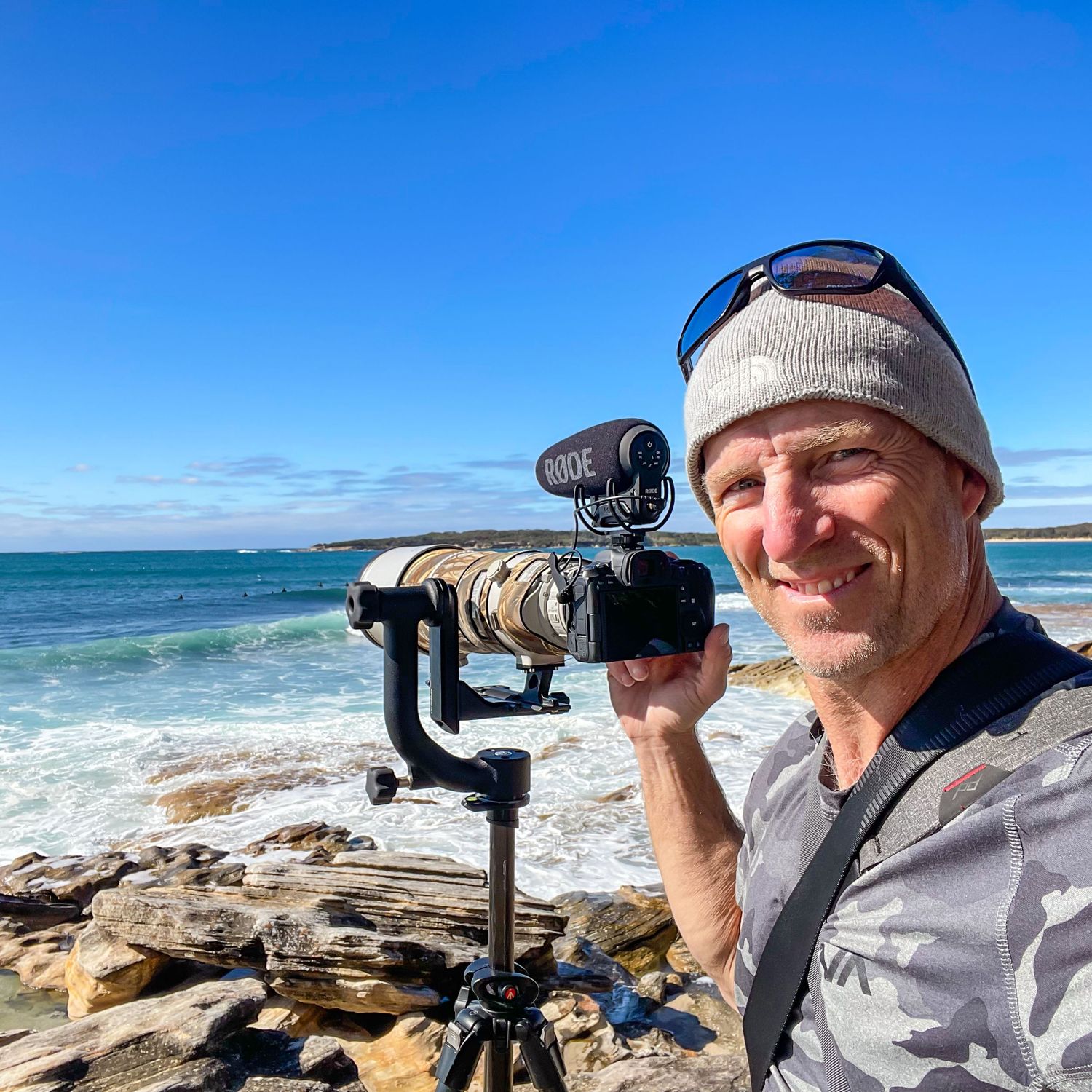 Nature photographer capturing coastal scenery with spotting scope and camera equipment on rocky beach overlook.
