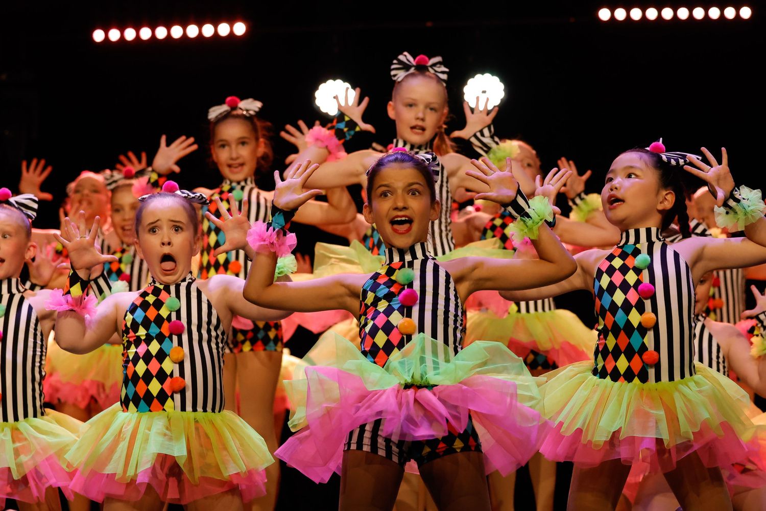 Dancers in colorful neon tutus and harlequin costumes perform a synchronized dance routine on stage.