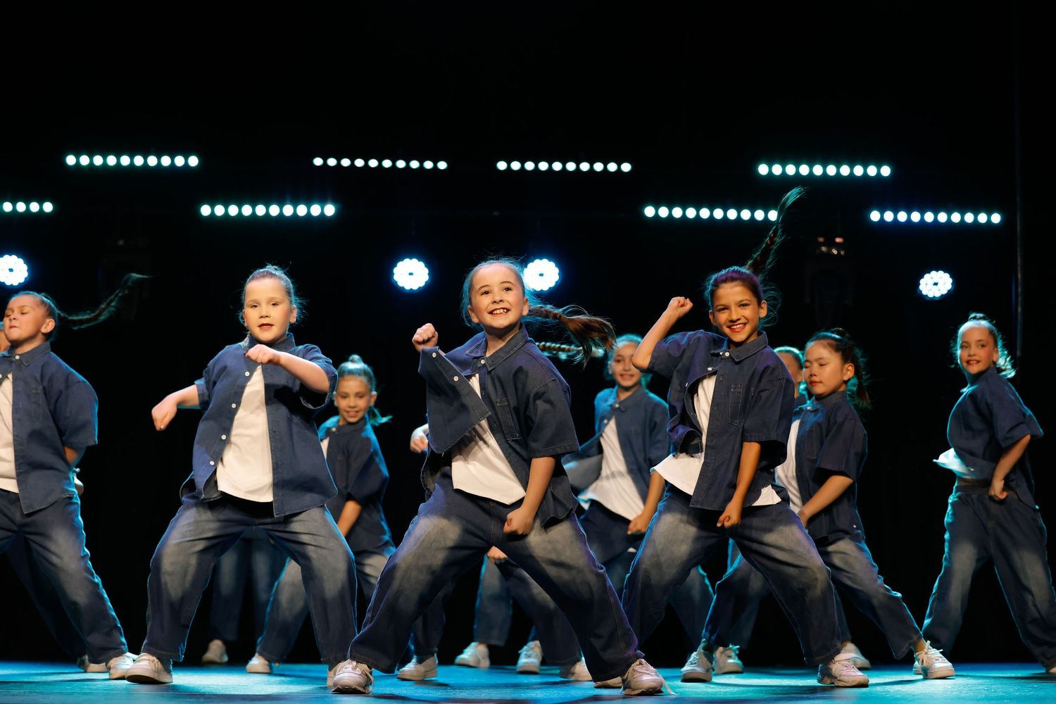 A synchronized dance performance on stage with performers in matching dark outfits creating a wave-like motion effect.