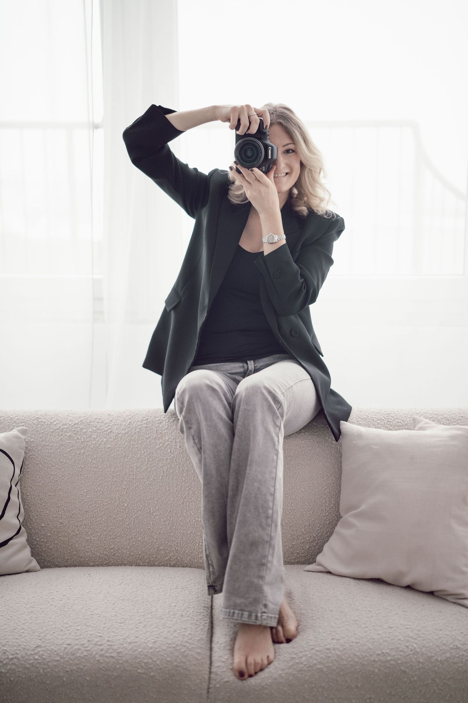 Photographer in black blazer and grey pants sits barefoot on beige couch while taking photo with digital camera.