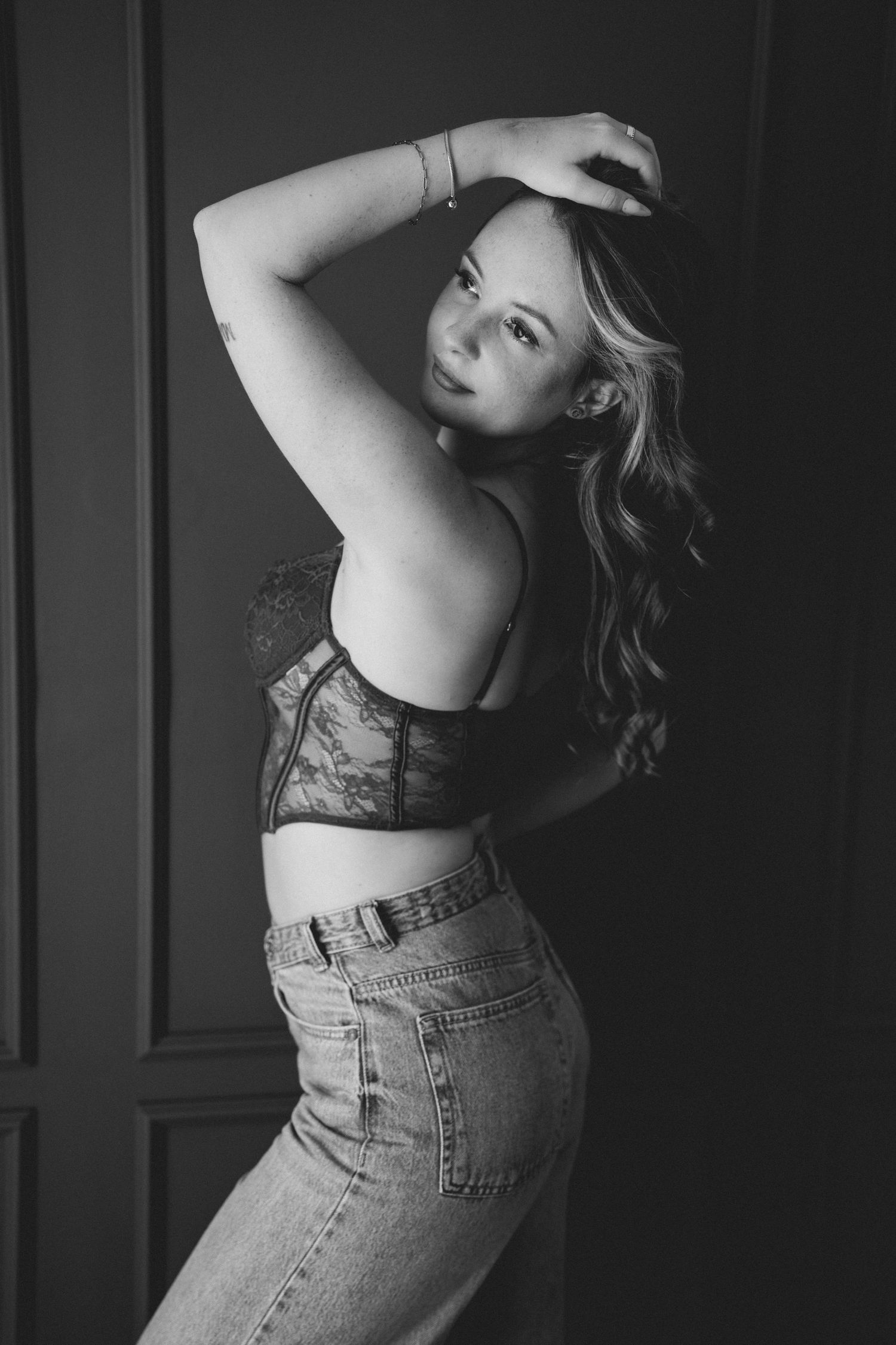 Black and white portrait of a young woman in lace bralette and jeans posing elegantly against a dark wall.
