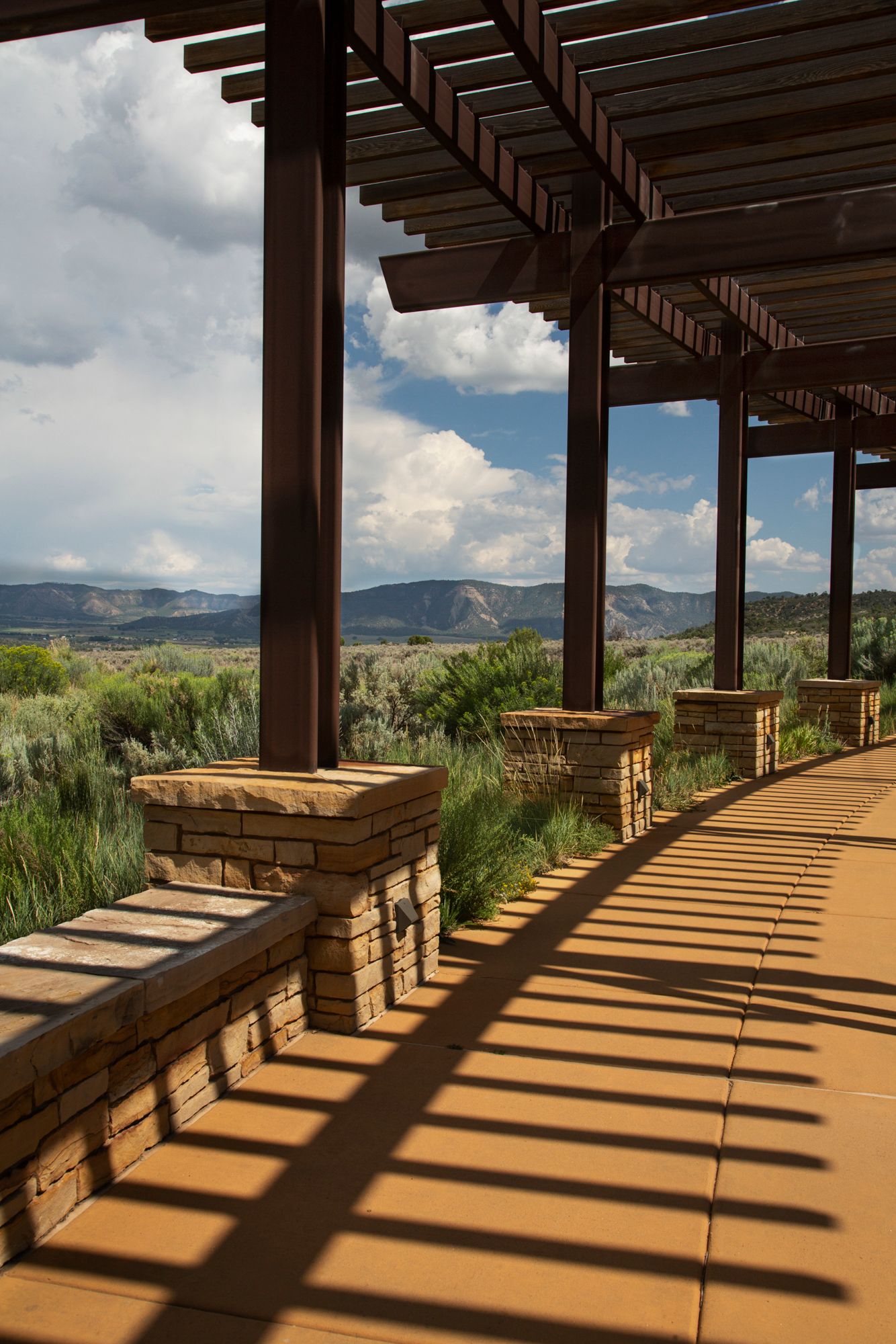 Covered patio walkway with wooden pergola and stone pillars casting dramatic shadows overlooking mountain landscape.