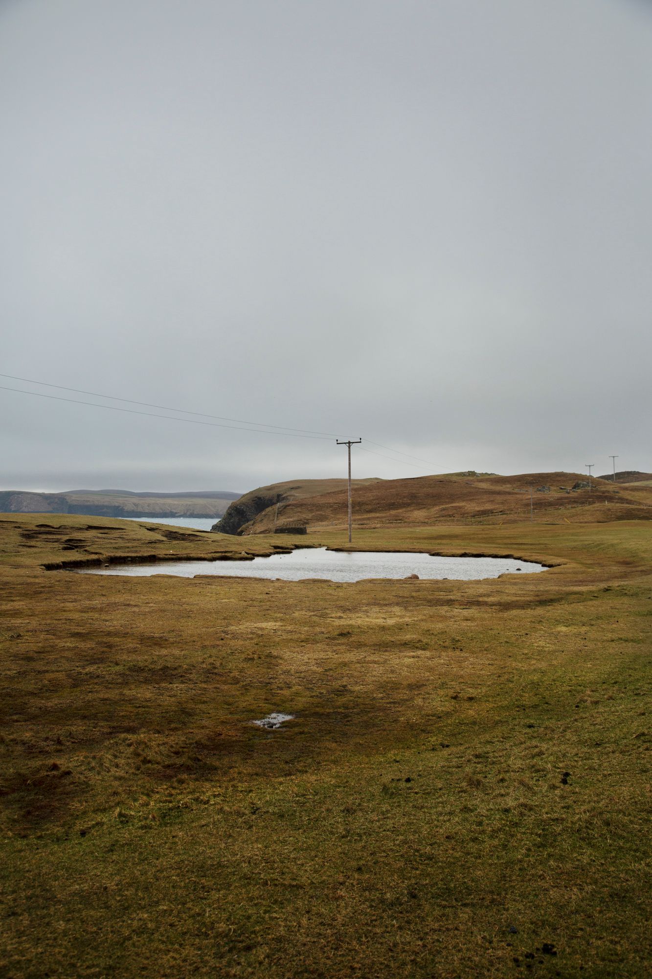 Rolling hills of green moorland with a small loch and utility poles stretching across the misty Scottish landscape.