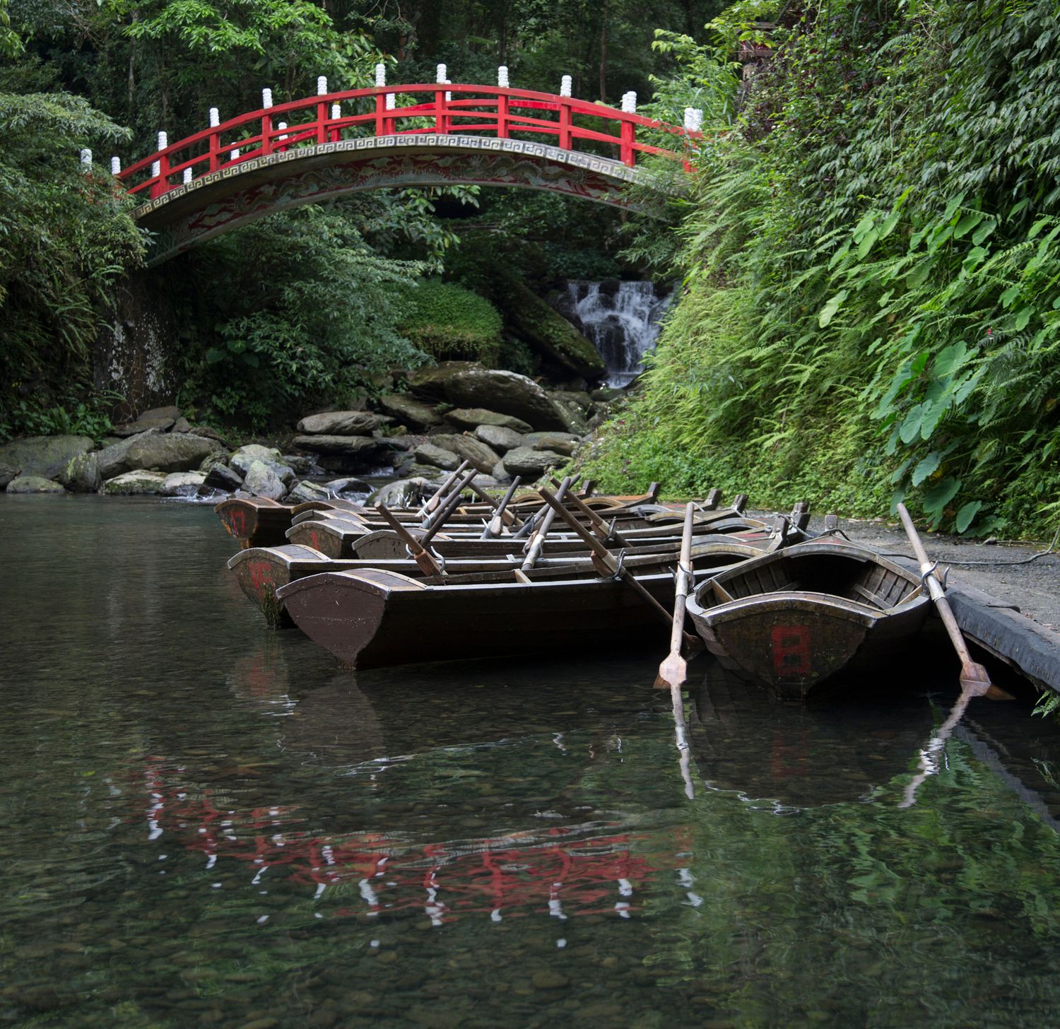 Red bridge spanning across dark water with lush greenery and rocks along shoreline.