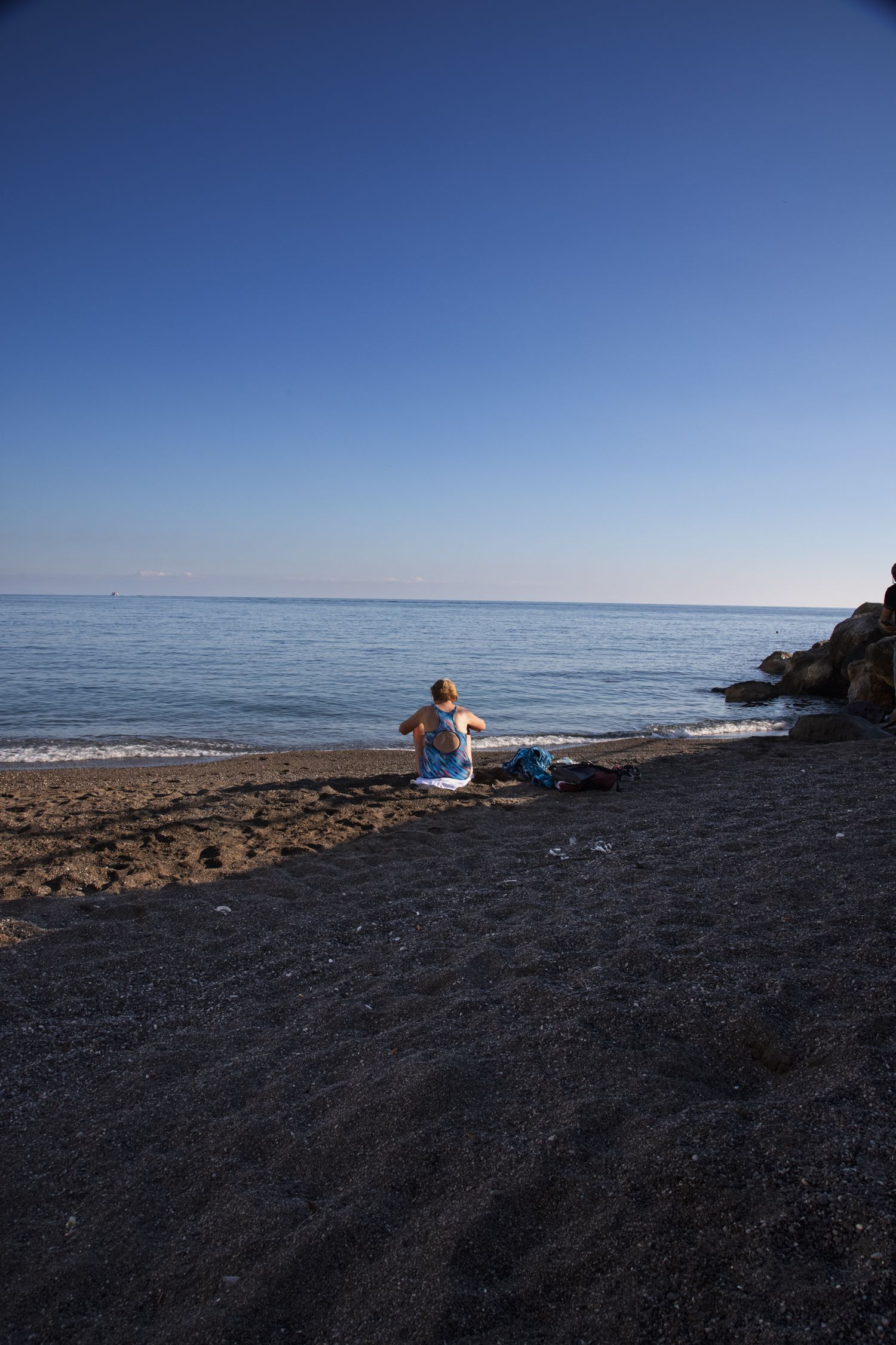 Person sitting alone on a dark sandy beach gazing at the calm blue sea under a clear sunny sky.