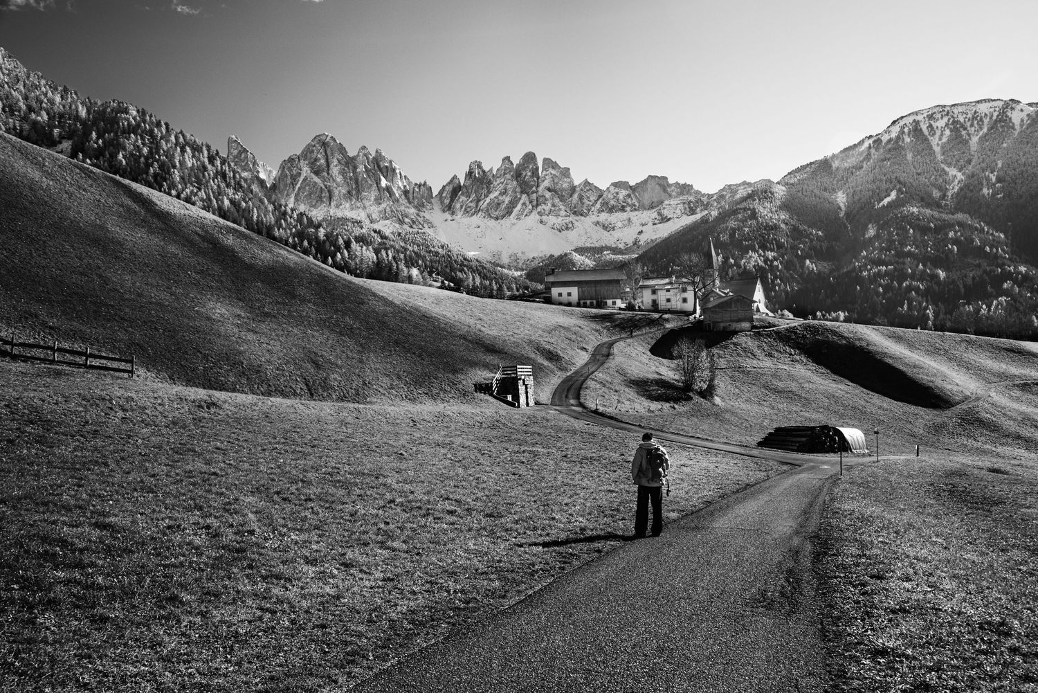 Black and white photo of a hiker on a mountain path leading to an alpine village with dramatic Dolomite peaks.