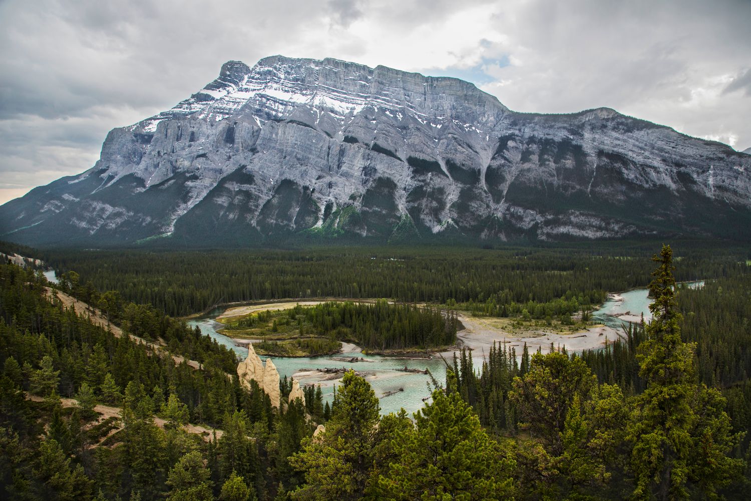 Mountain landscape with snow-capped peaks and winding river through forested valley.