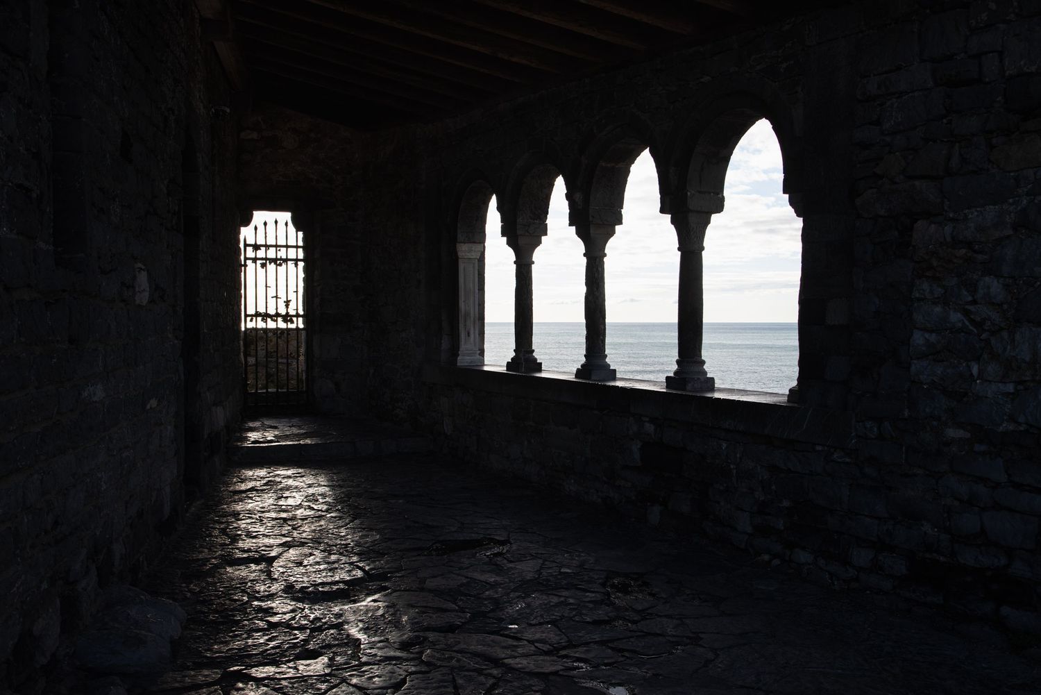 Dark stone archway colonnade with Gothic arches framing a serene ocean view in dramatic silhouette lighting.