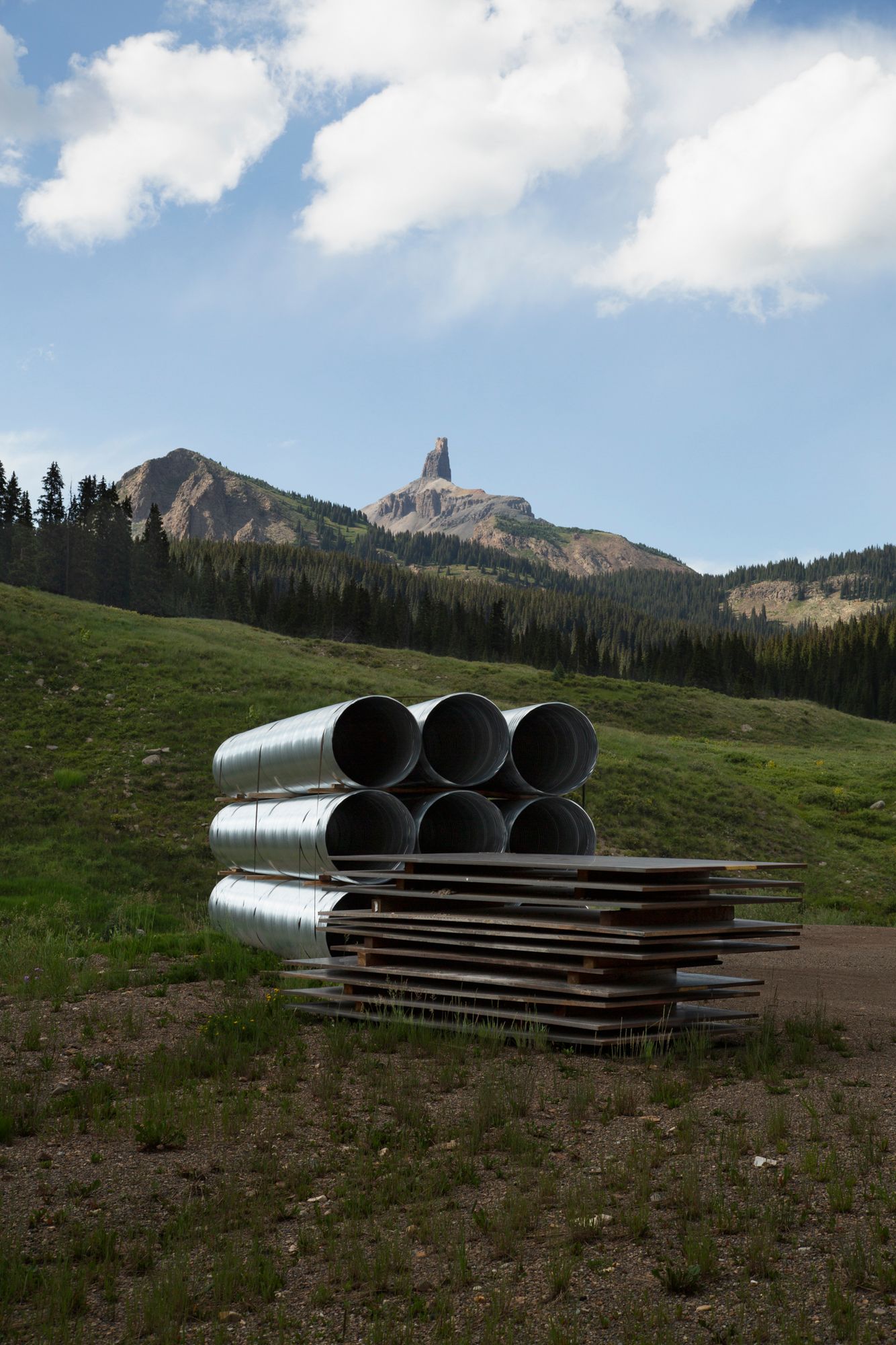 Large metal pipes are stacked on a grassy mountainside with rugged peaks and blue sky in the background.