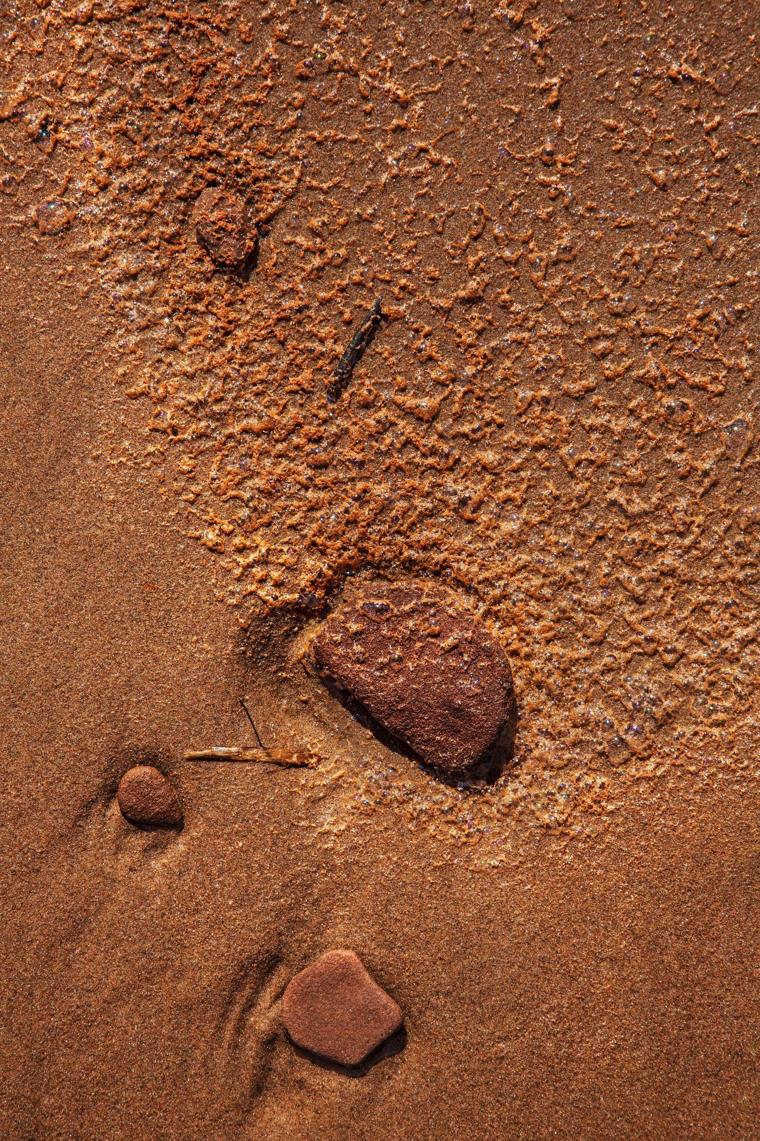 Close-up of rust-colored sandy beach with small stones and pebbles creating natural patterns in wet red sand.
