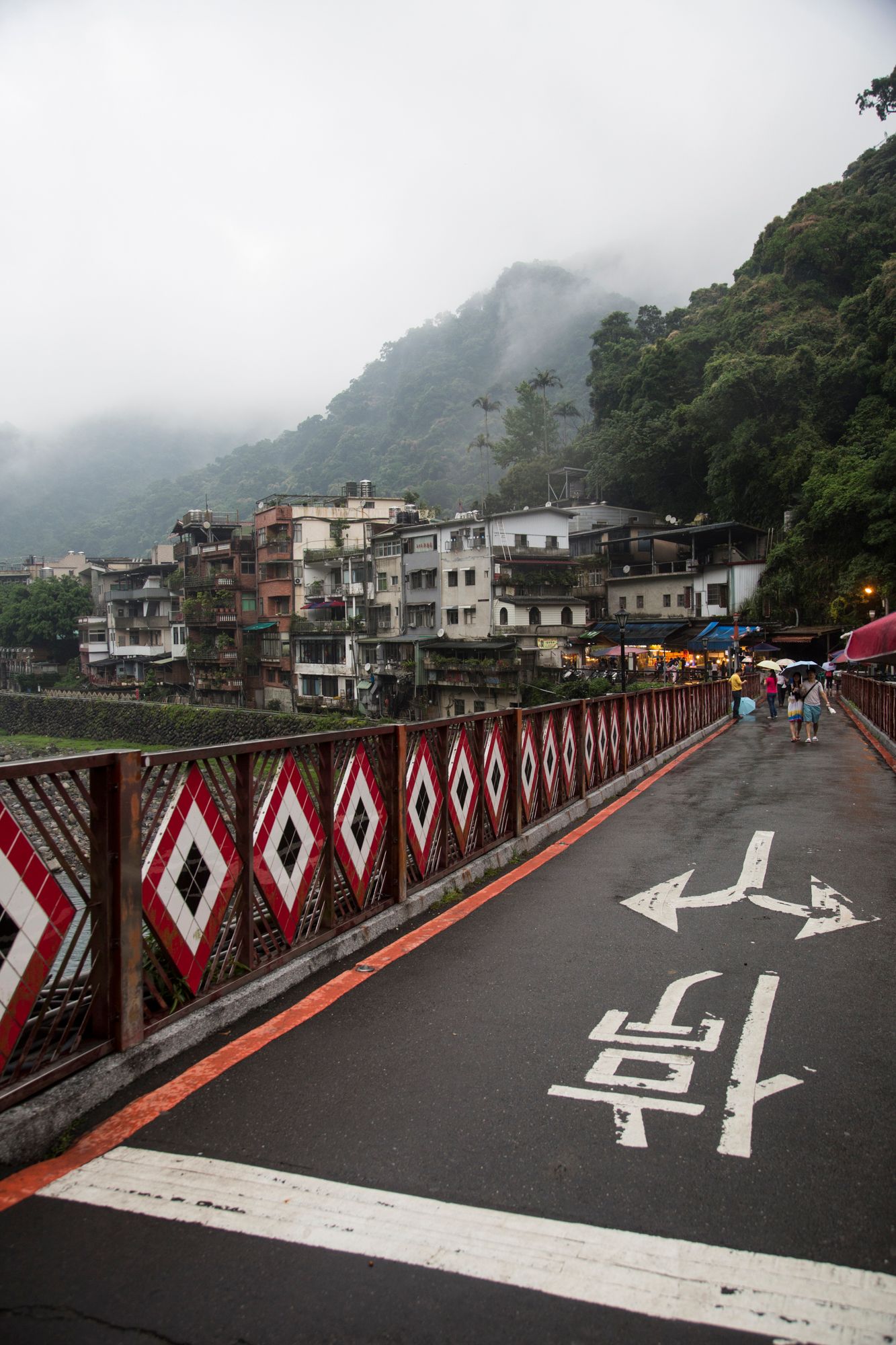 Bridge with red and white diamond pattern railings leading to mountain village.