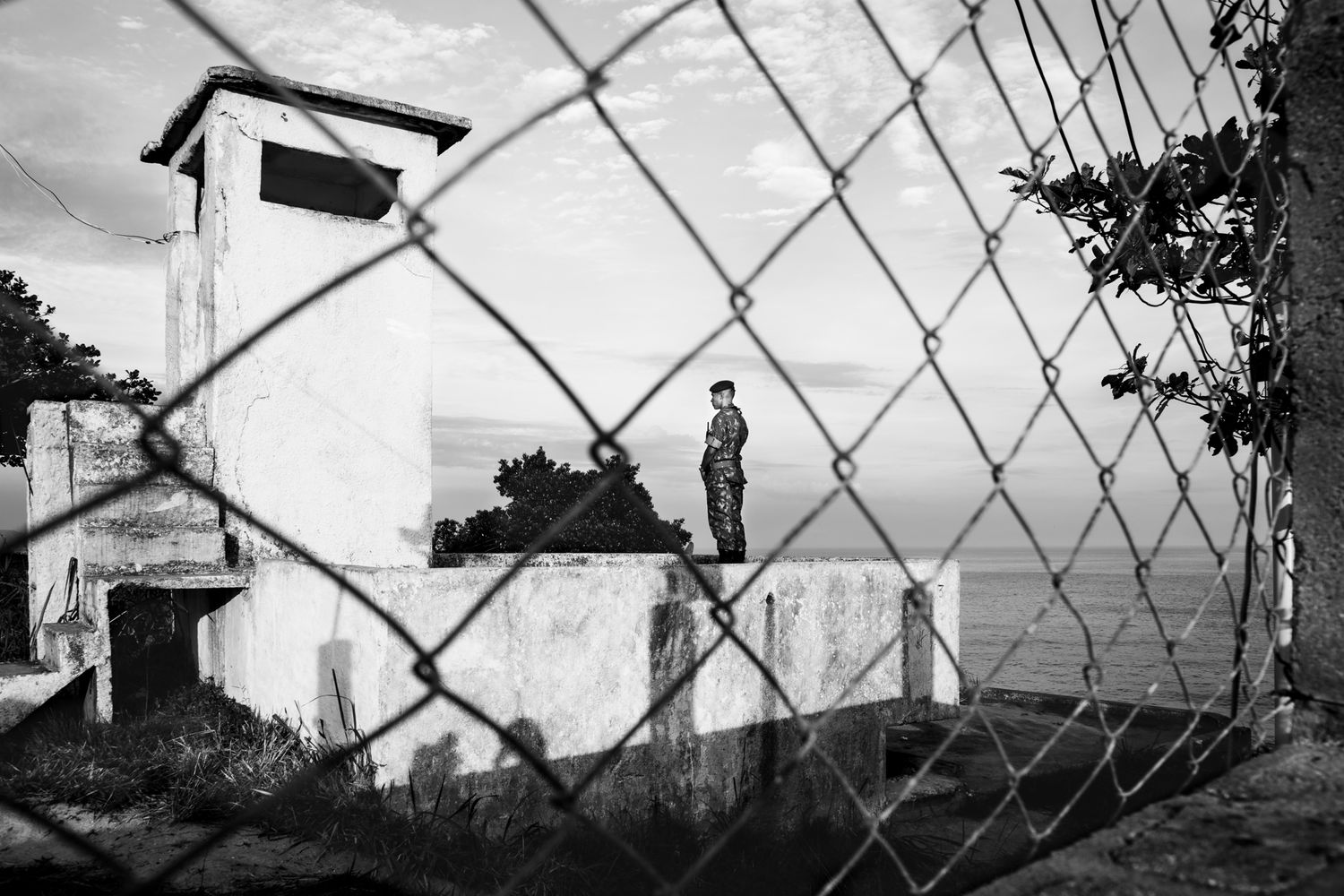 Urban skyline viewed through chain-link fence with industrial structures in black and white.