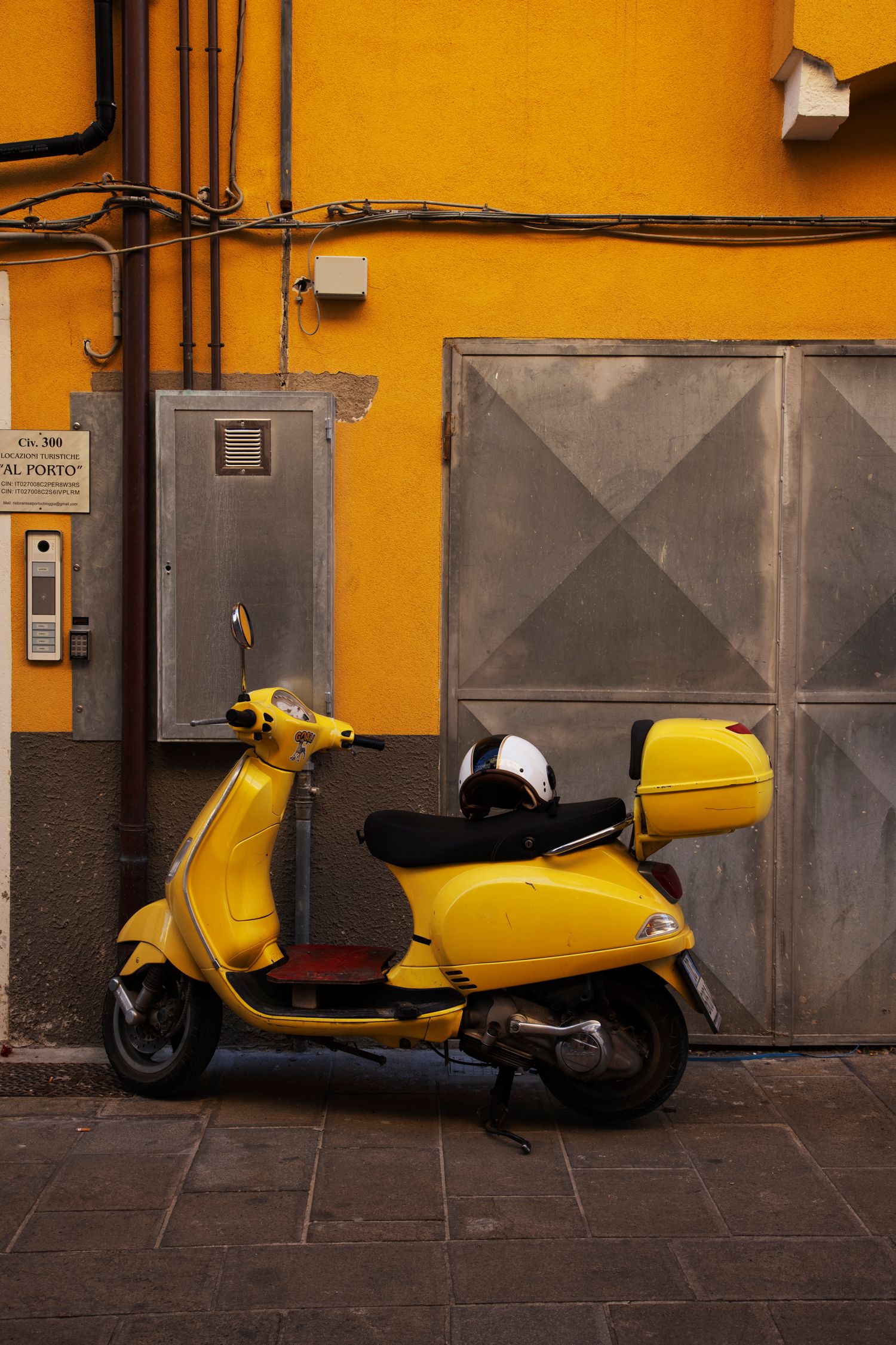 Yellow Vespa scooter parked against a vibrant orange wall with metal garage door in an Italian street.