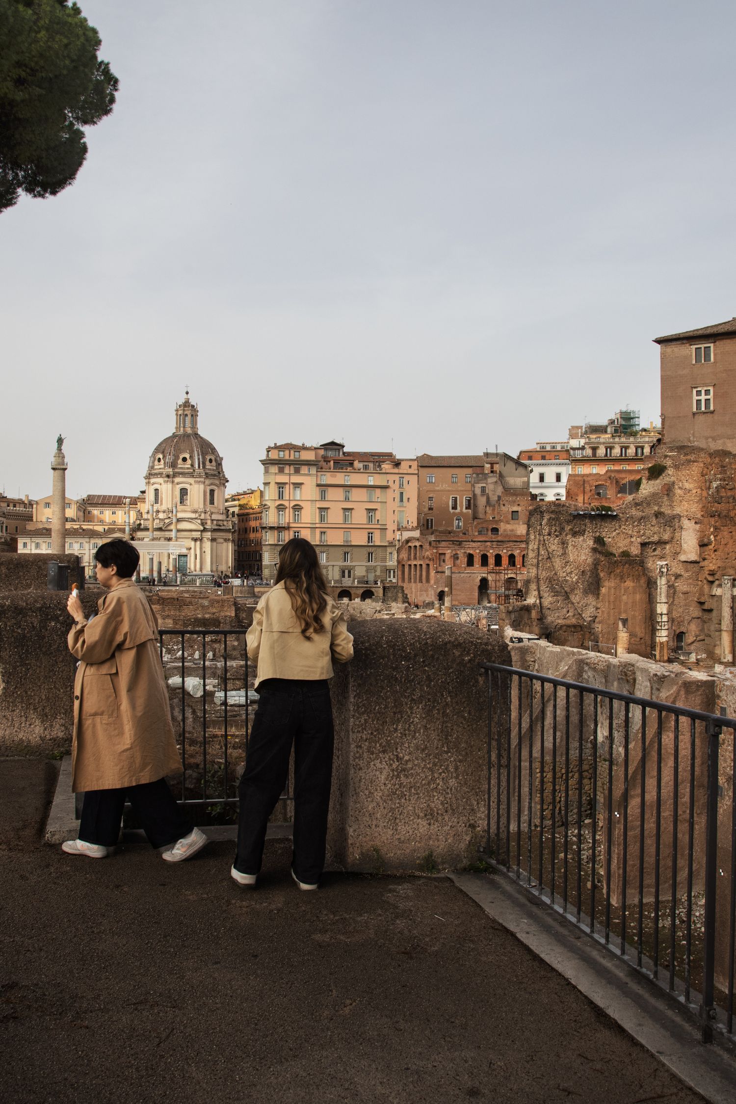 Two women overlook ancient Roman ruins and domed church from a terrace viewpoint in Rome, Italy.