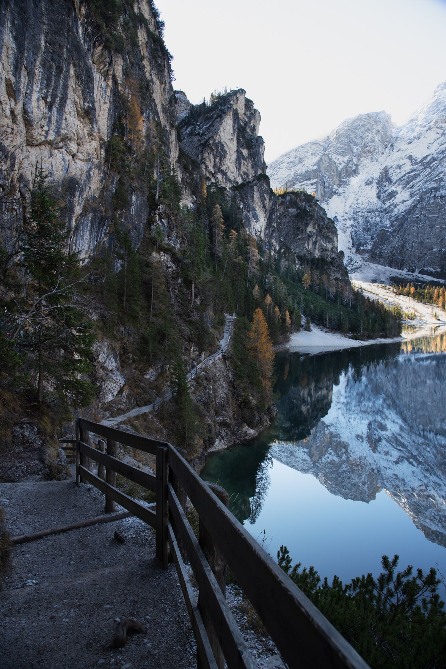 Wooden railing path along a reflective alpine lake with towering rocky cliffs and snow-dusted evergreen trees.