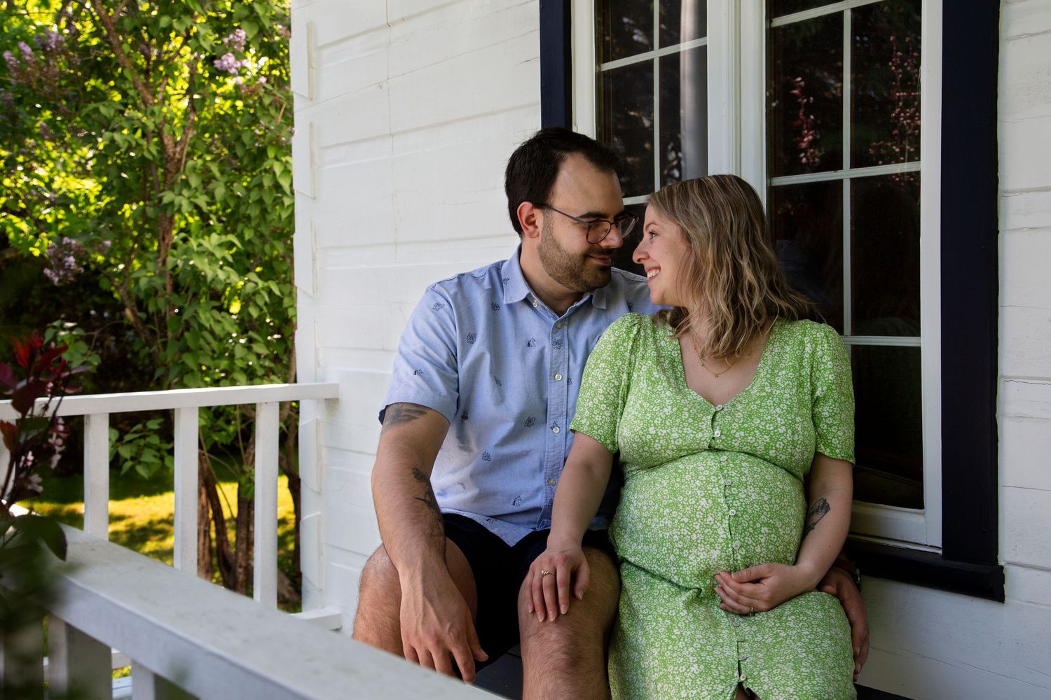 Outdoor maternity photos on porch with couple in casual attire against white house exterior.