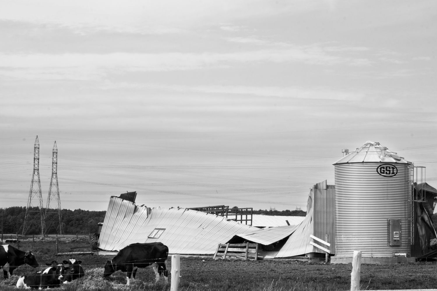Industrial storage tanks and power lines stand against a cloudy sky in a panoramic black and white landscape view.