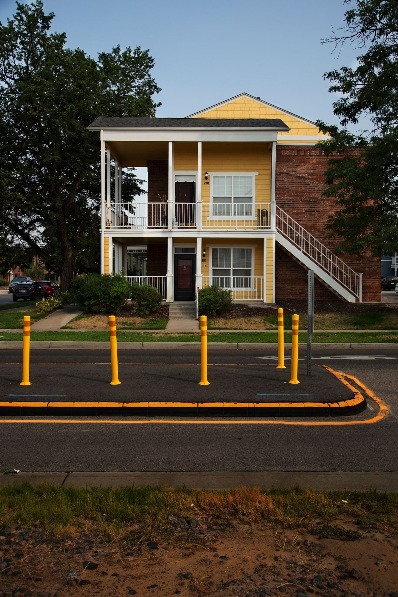 Two-story apartment building with yellow siding, white columns, and a covered balcony behind yellow traffic posts.