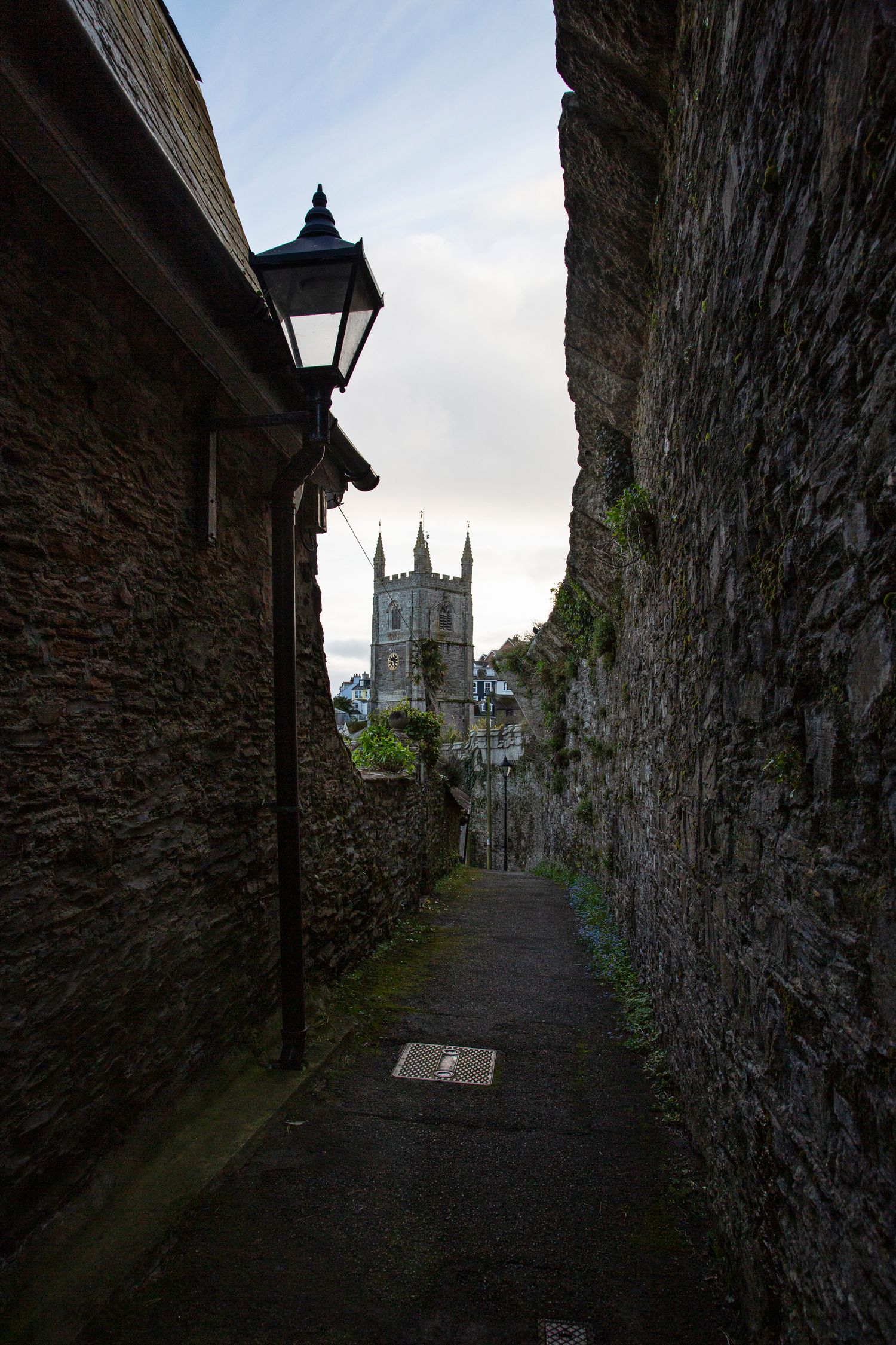 Narrow stone alleyway with traditional lamp post leading to a church tower in the distance.