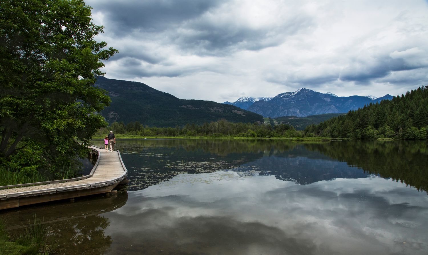 Wooden boardwalk extends over calm mountain lake reflecting cloudy skies and distant peaks.