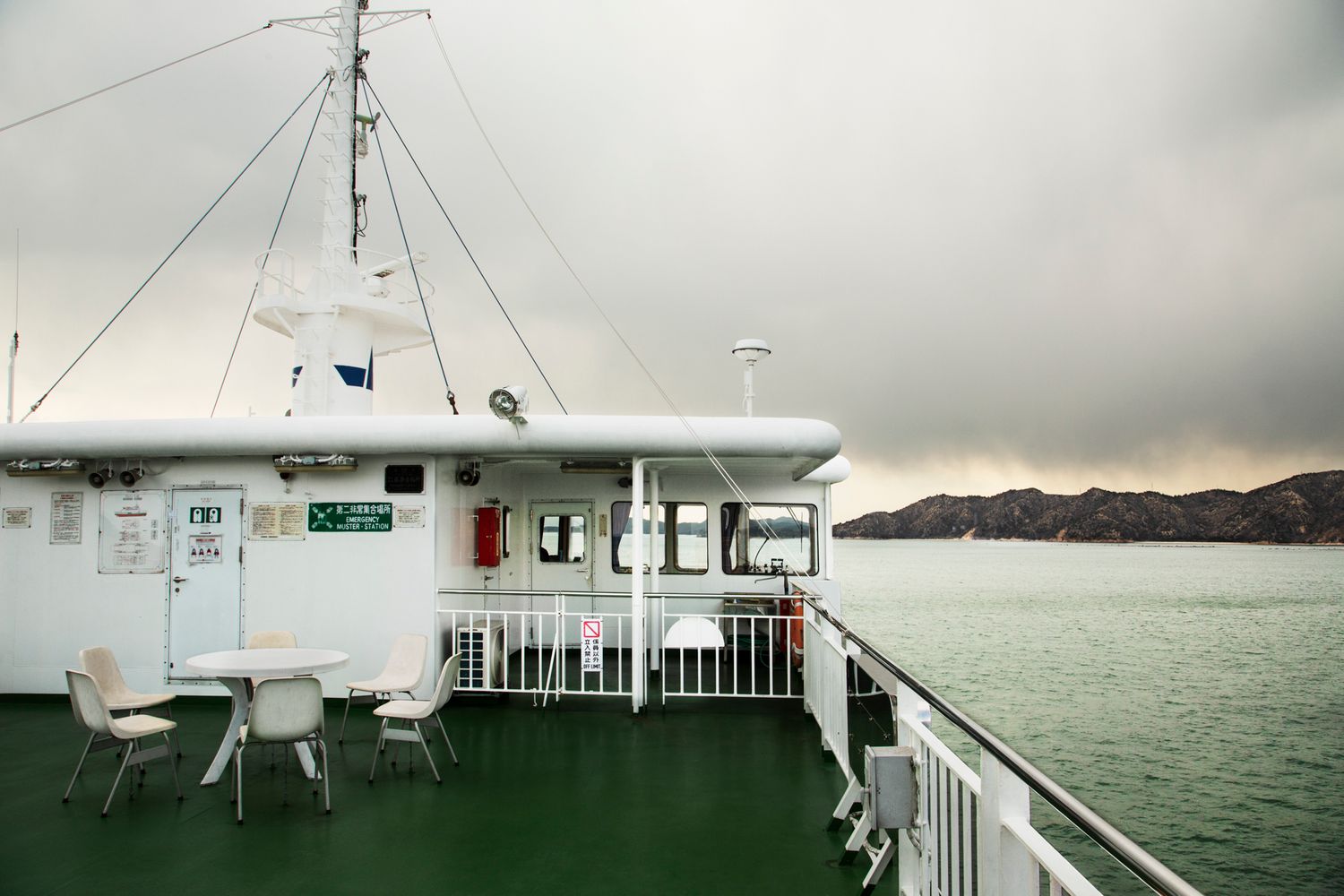 Panoramic view from a ferry deck showing rails, seating, and distant mountains across the water.