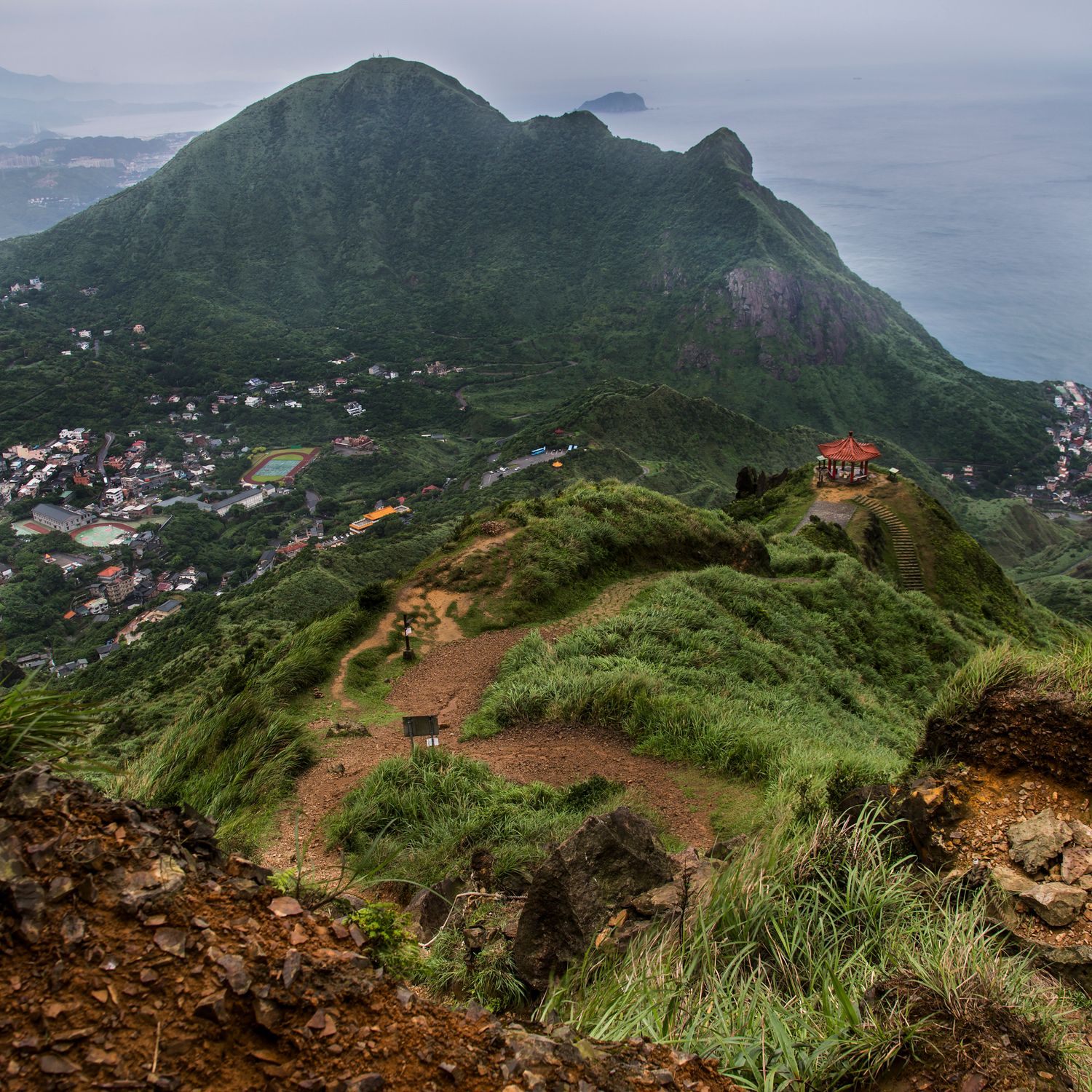 Winding mountain trail overlooking coastal settlements with lush green slopes and distant ocean views.