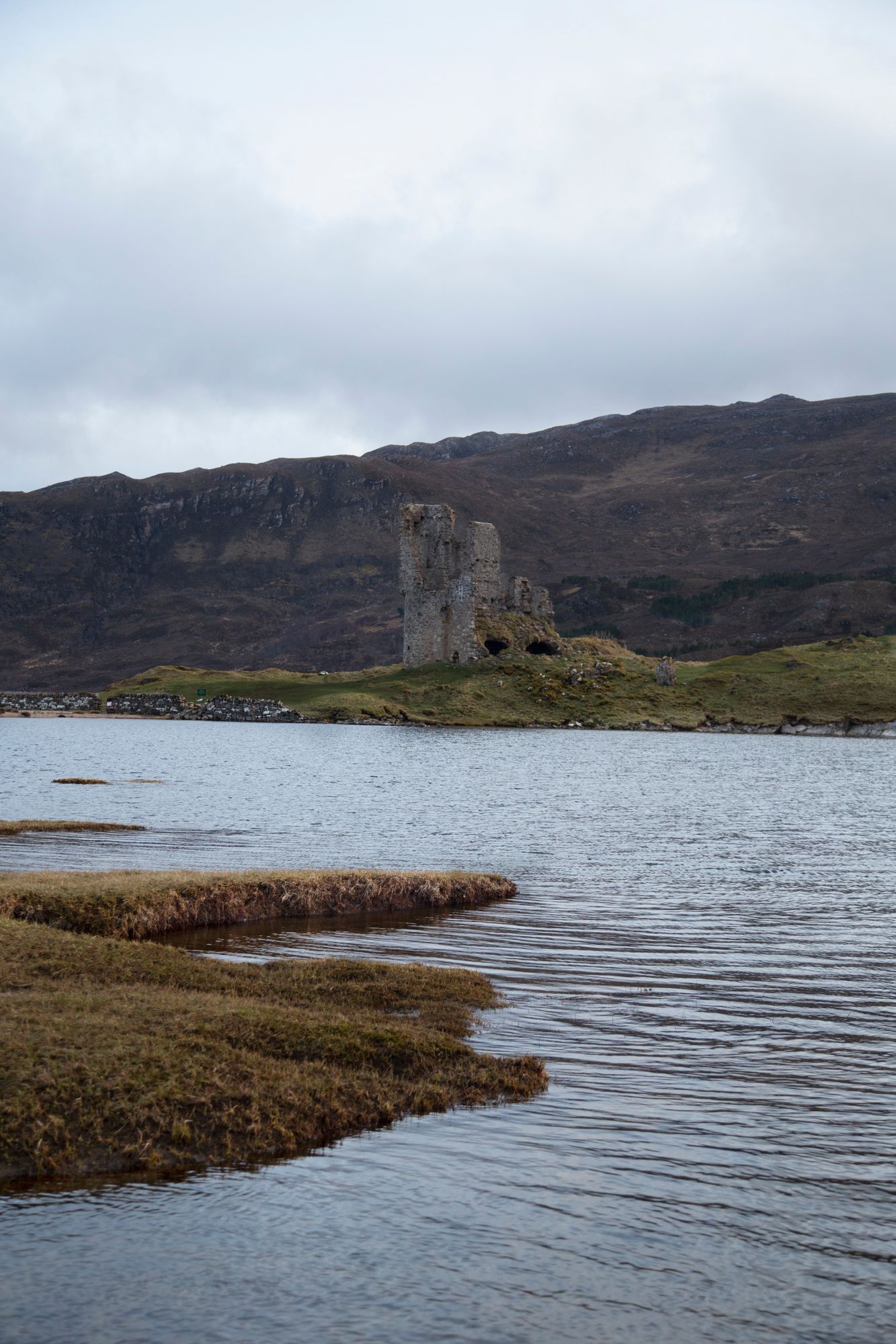 Panoramic view of a rugged Scottish coastline with castle ruins visible on the cliffs overlooking a tranquil loch.