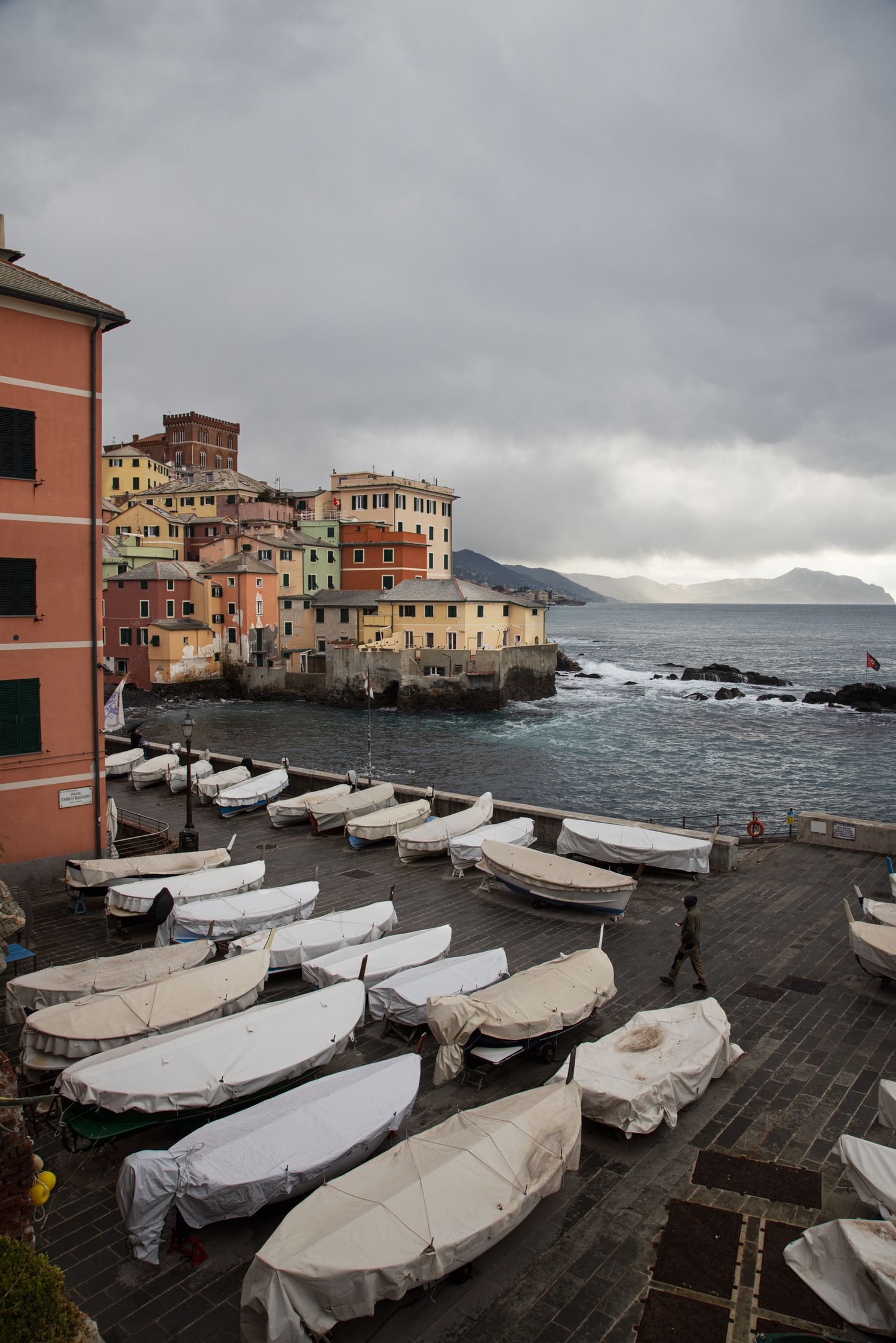 Colorful Italian coastal village with white-covered boats on a waterfront plaza under dramatic cloudy skies.