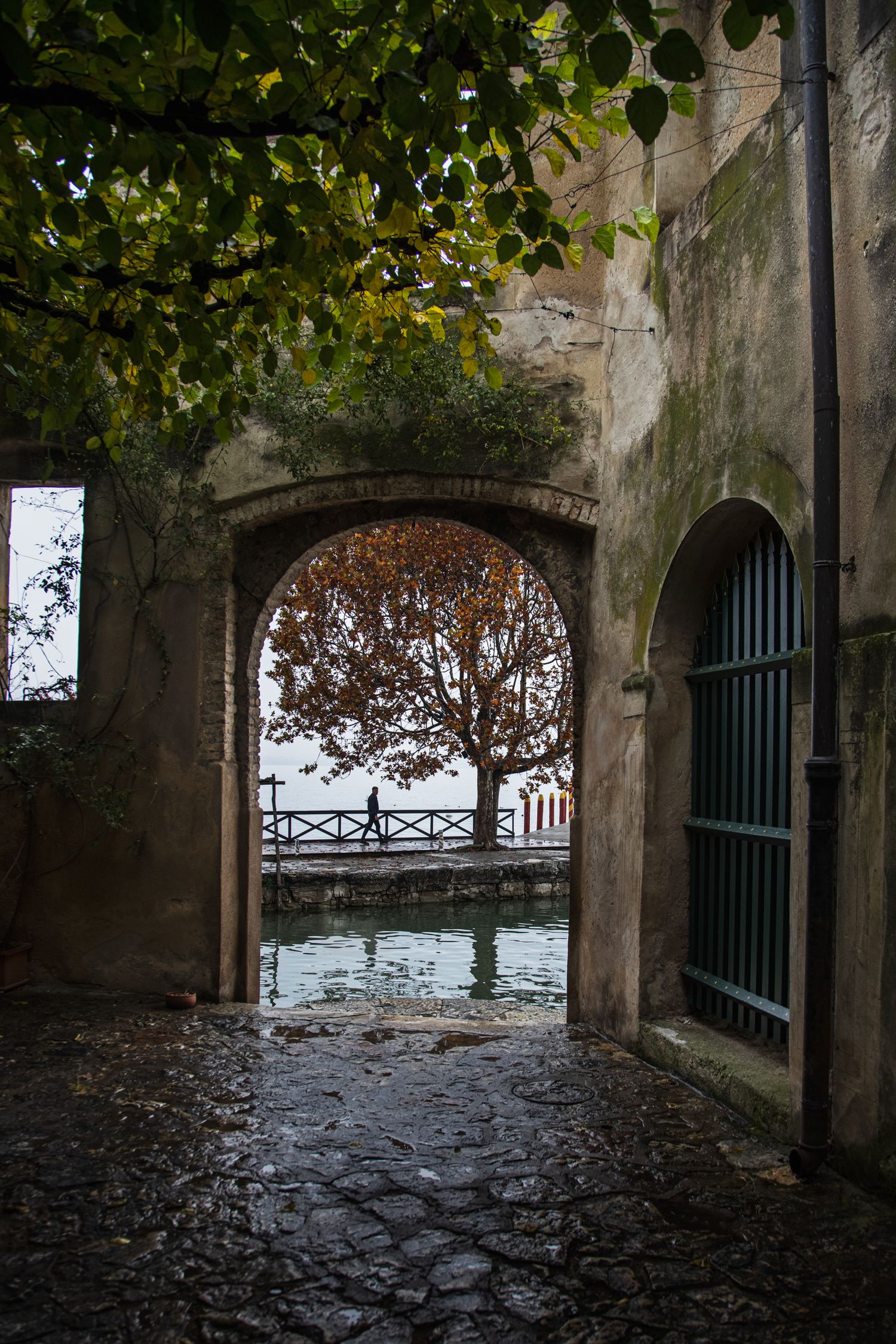 Stone archway framing autumn tree and waterfront in an ancient Italian courtyard with cobblestone path.