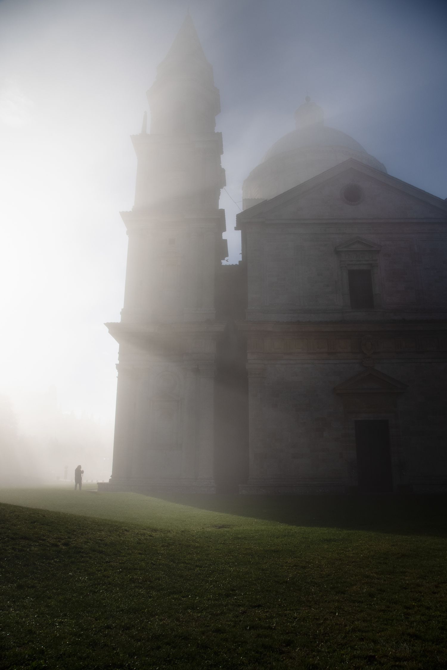 Misty foggy church with bell tower on green hill, lone figure standing beside ancient stone cathedral in atmospheric fog.