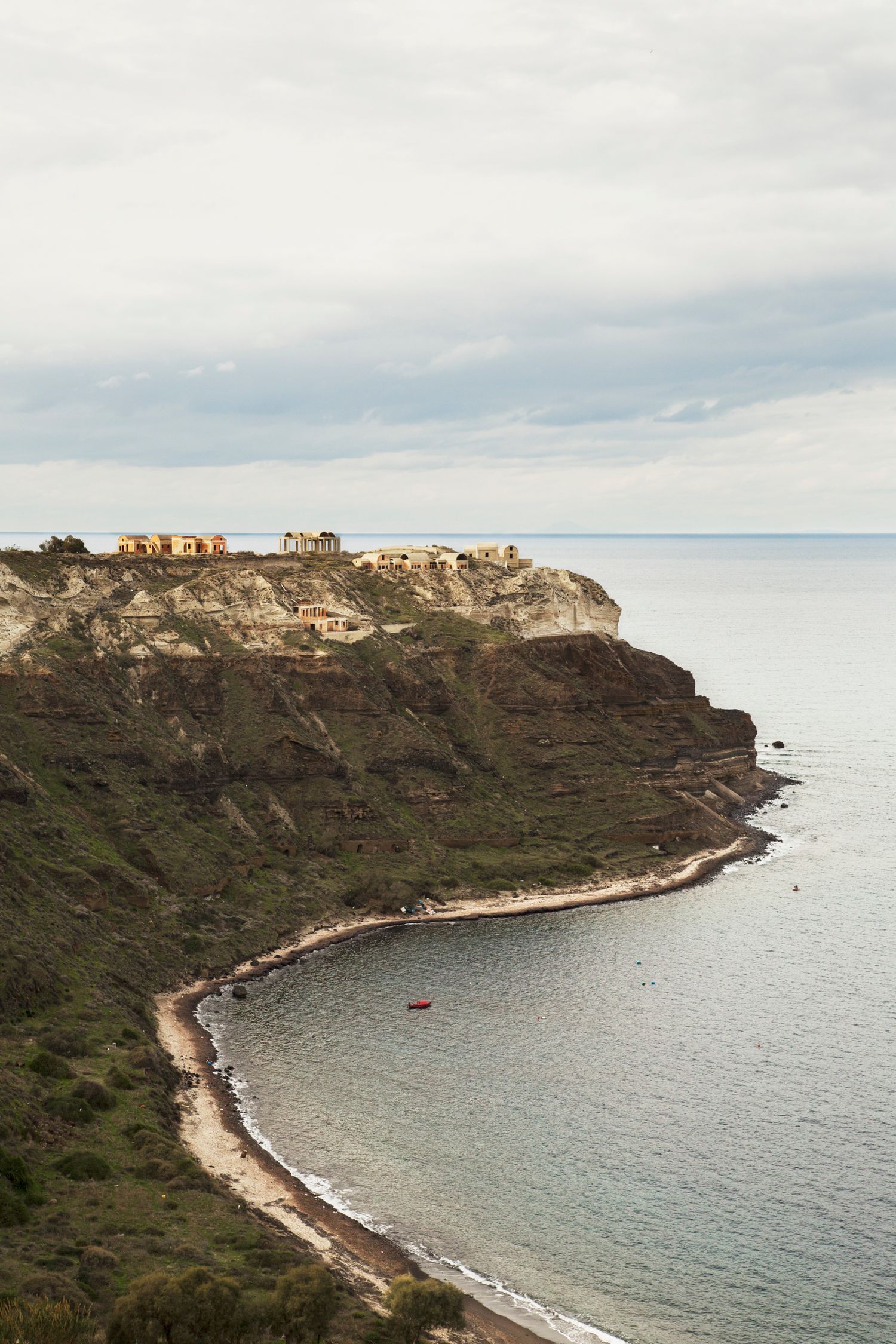 Buildings perched on steep coastal cliffs overlooking a curved shoreline and calm Mediterranean waters.