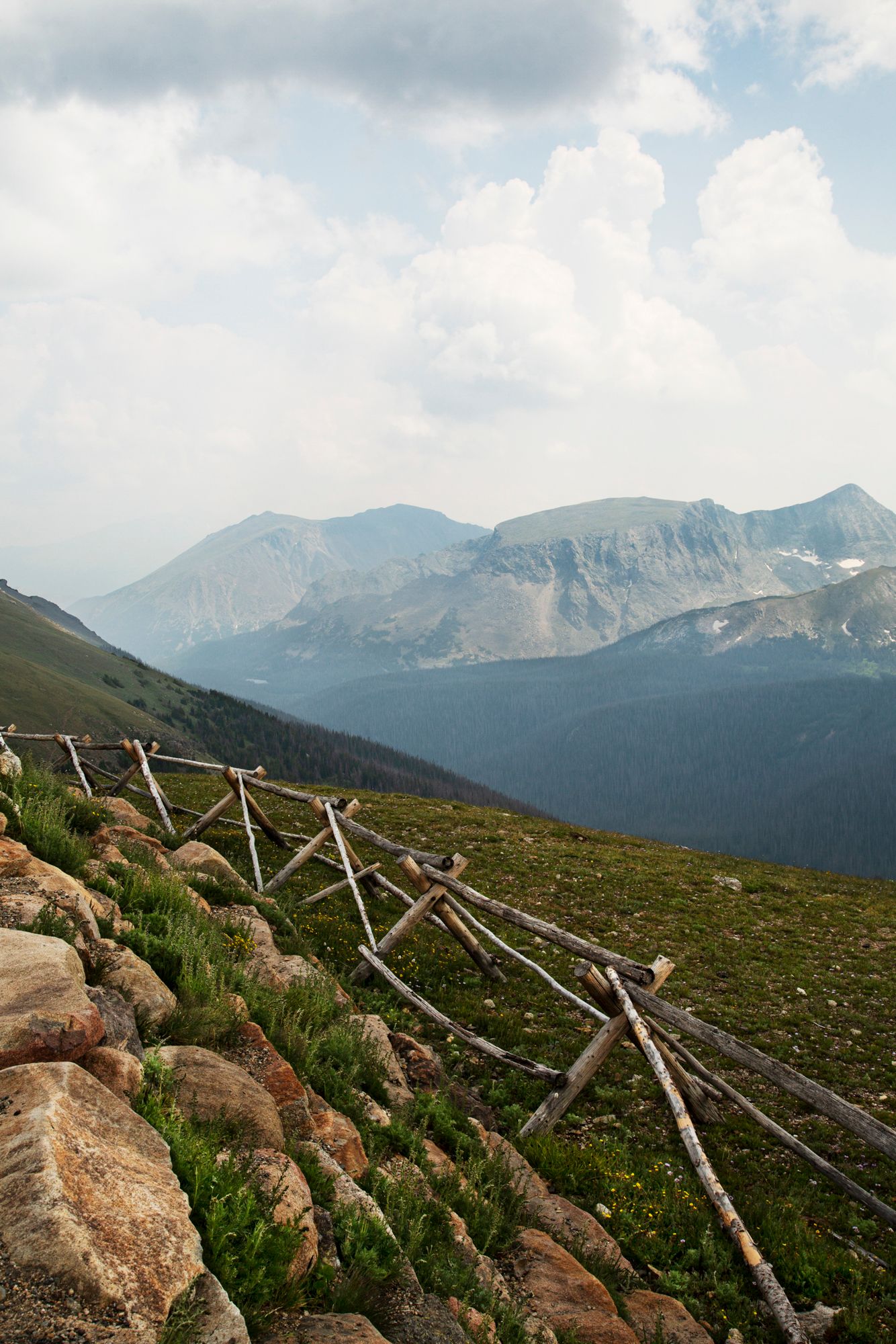 A wooden split-rail fence runs along a rocky mountain trail with majestic peaks and cloudy skies in the background.