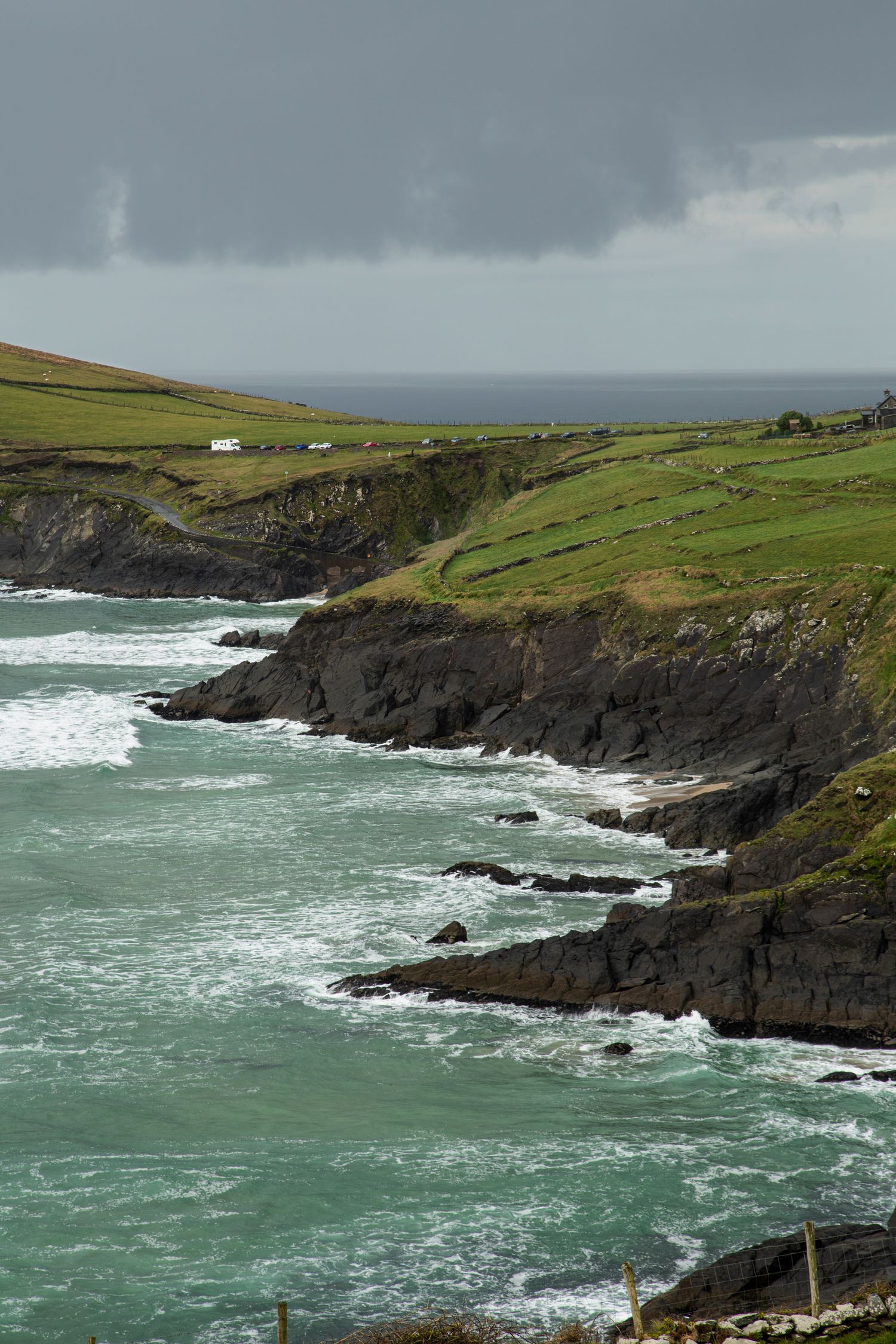 Rugged coastline with crashing waves against dark cliffs and green fields overlooking the sea.