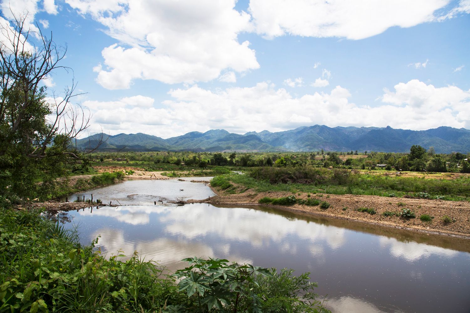 Wide river landscape with clouds reflected in calm water and mountains in the distance.