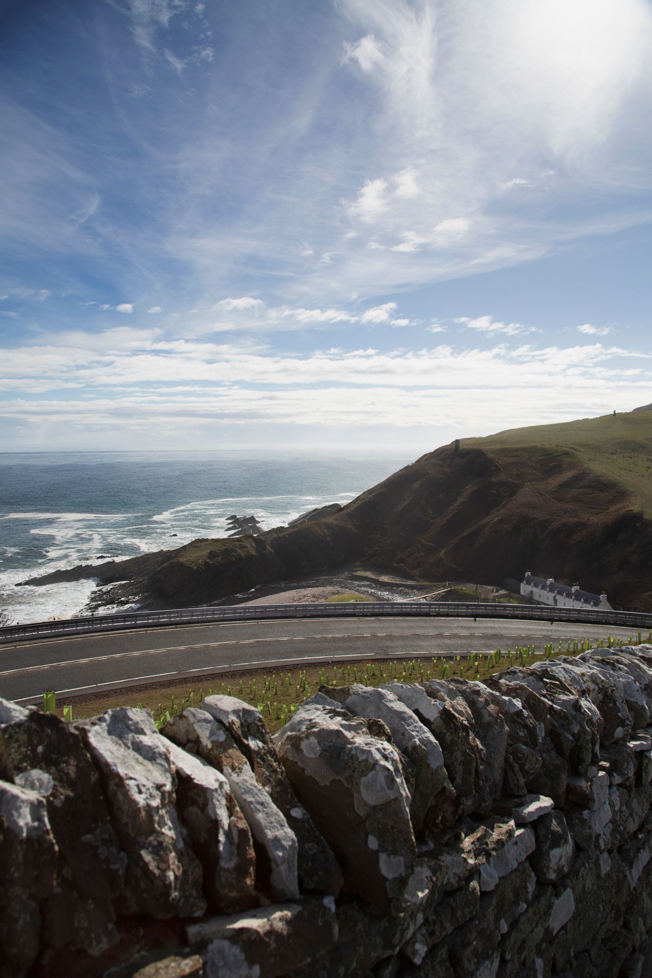 Panoramic coastal view showing rugged cliffs, crashing waves, and a winding road along the Pacific Ocean under cloudy skies.