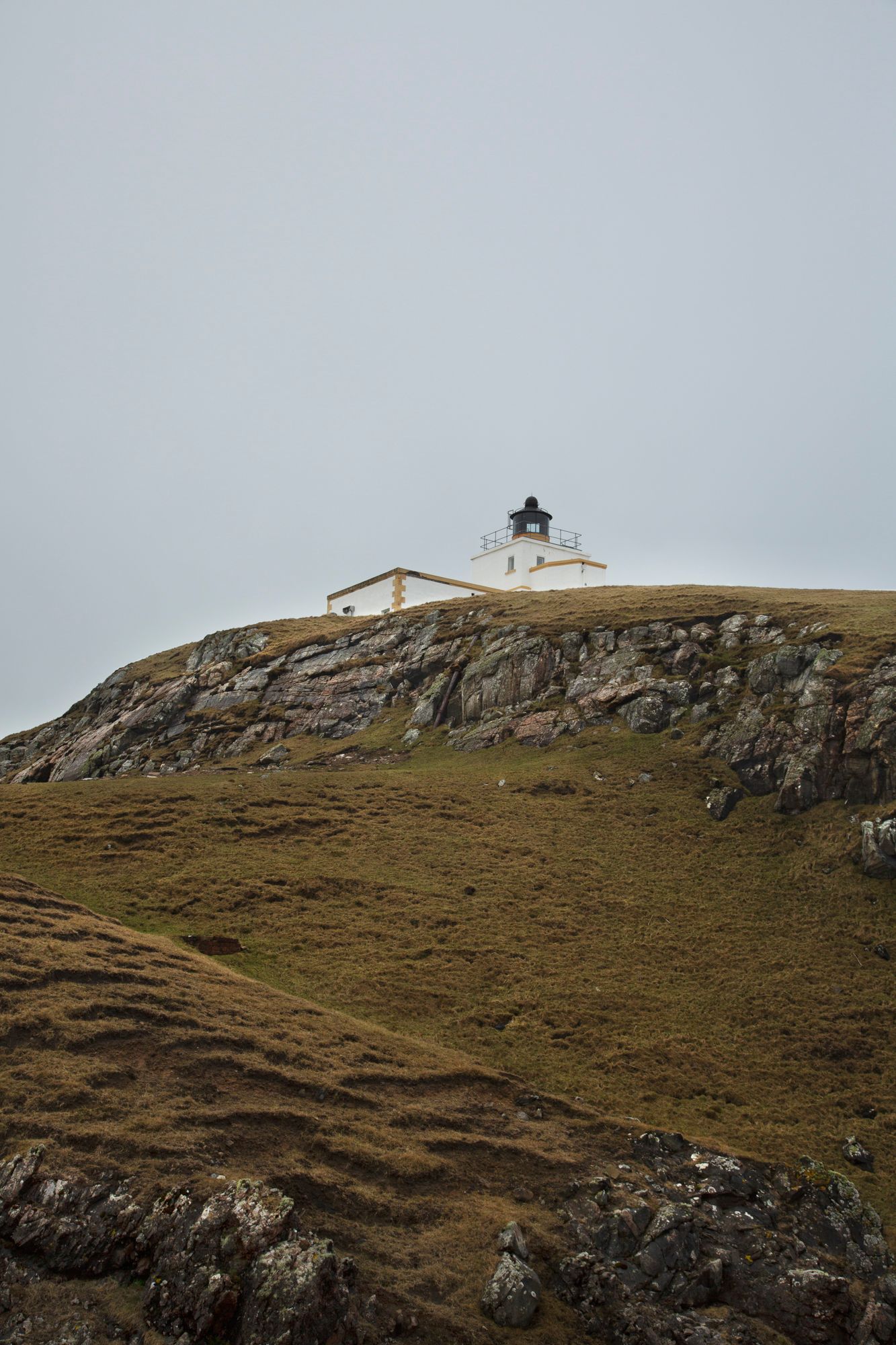 A white lighthouse perches atop a rugged coastal cliff with grassy slopes and rocky outcrops in the Scottish highlands.
