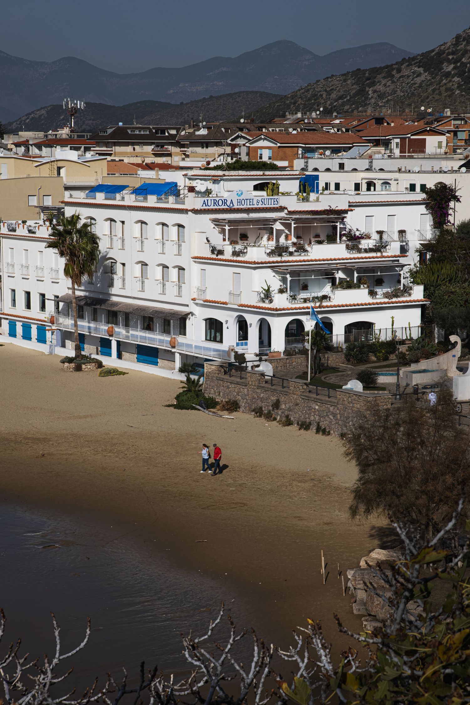 Aerial view of Hotel Aurora on a sandy beach in a coastal Mediterranean town with mountains in the background.