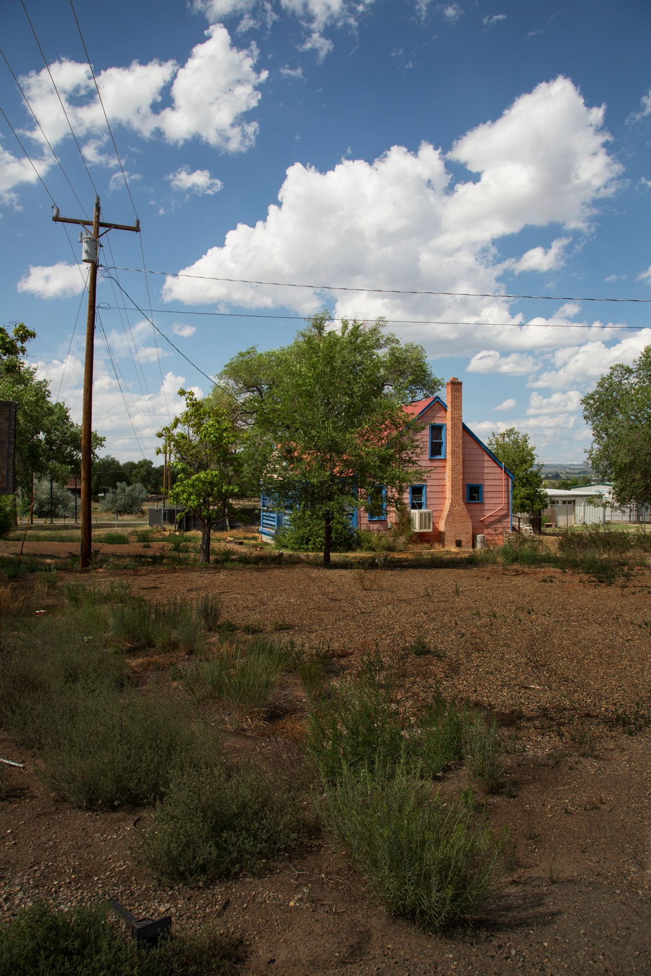 A panoramic view of a rural property with a brick house, utility poles, and scattered vegetation under a cloudy blue sky.