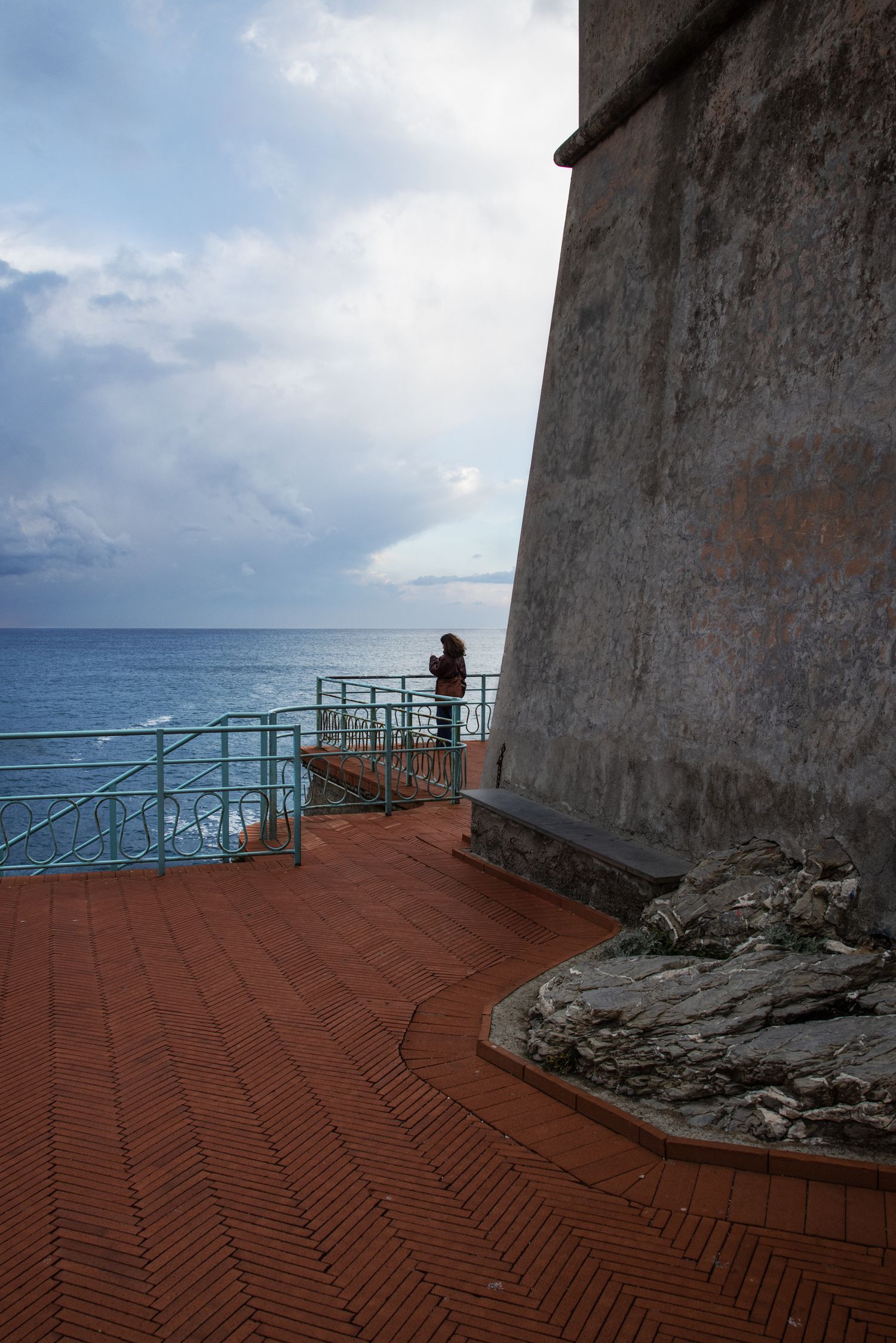 Person standing on a terrace by the sea wall overlooking the blue ocean under a cloudy sky.
